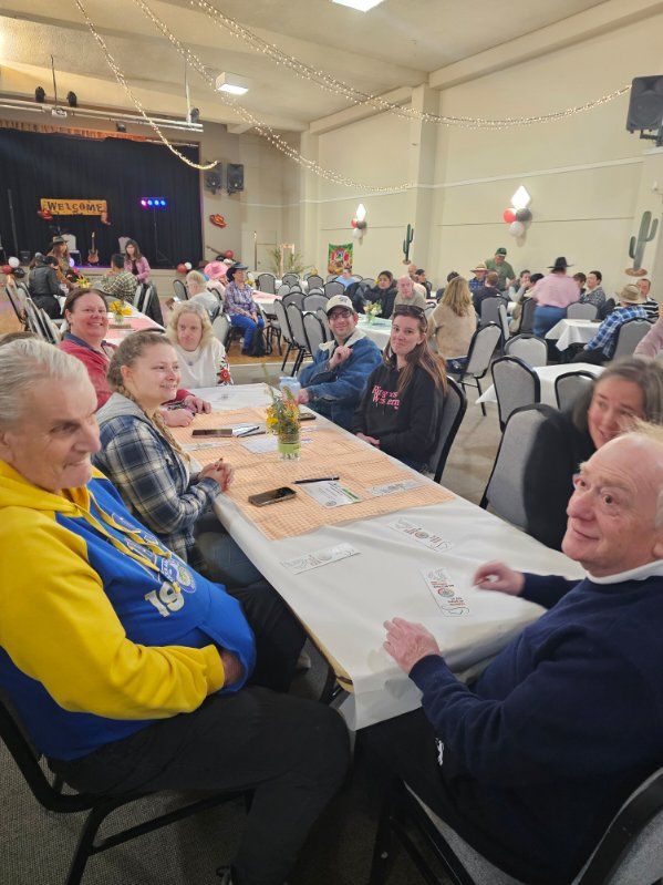 People at tables in a brightly lit hall, some playing dominoes, others conversing. Stage in background.