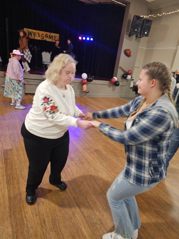 Two women dancing in a hall; one with Down syndrome, smiling. Others in the background near a stage.