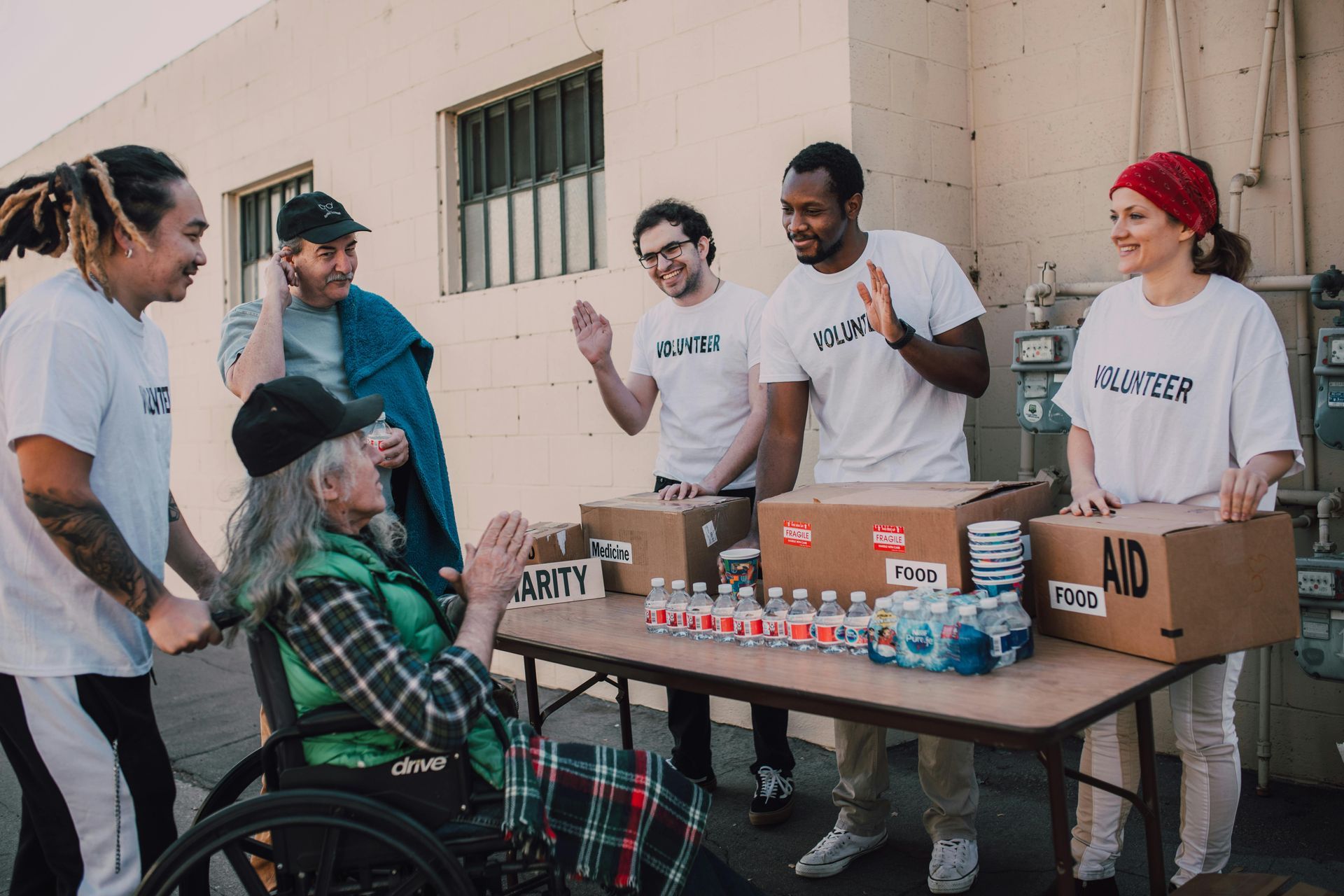 Volunteers giving supplies to a person in a wheelchair. Donations on table outside.