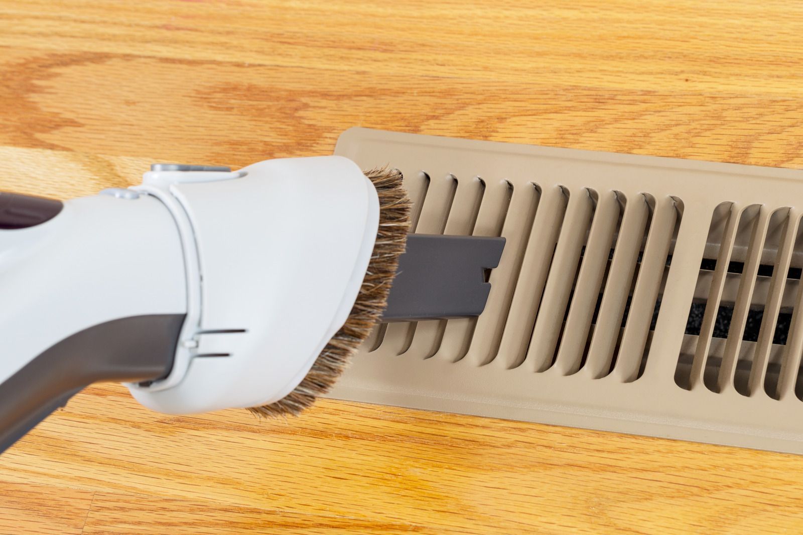 A vacuum brush attachment cleaning dust from a beige floor vent installed in wood-grain flooring.