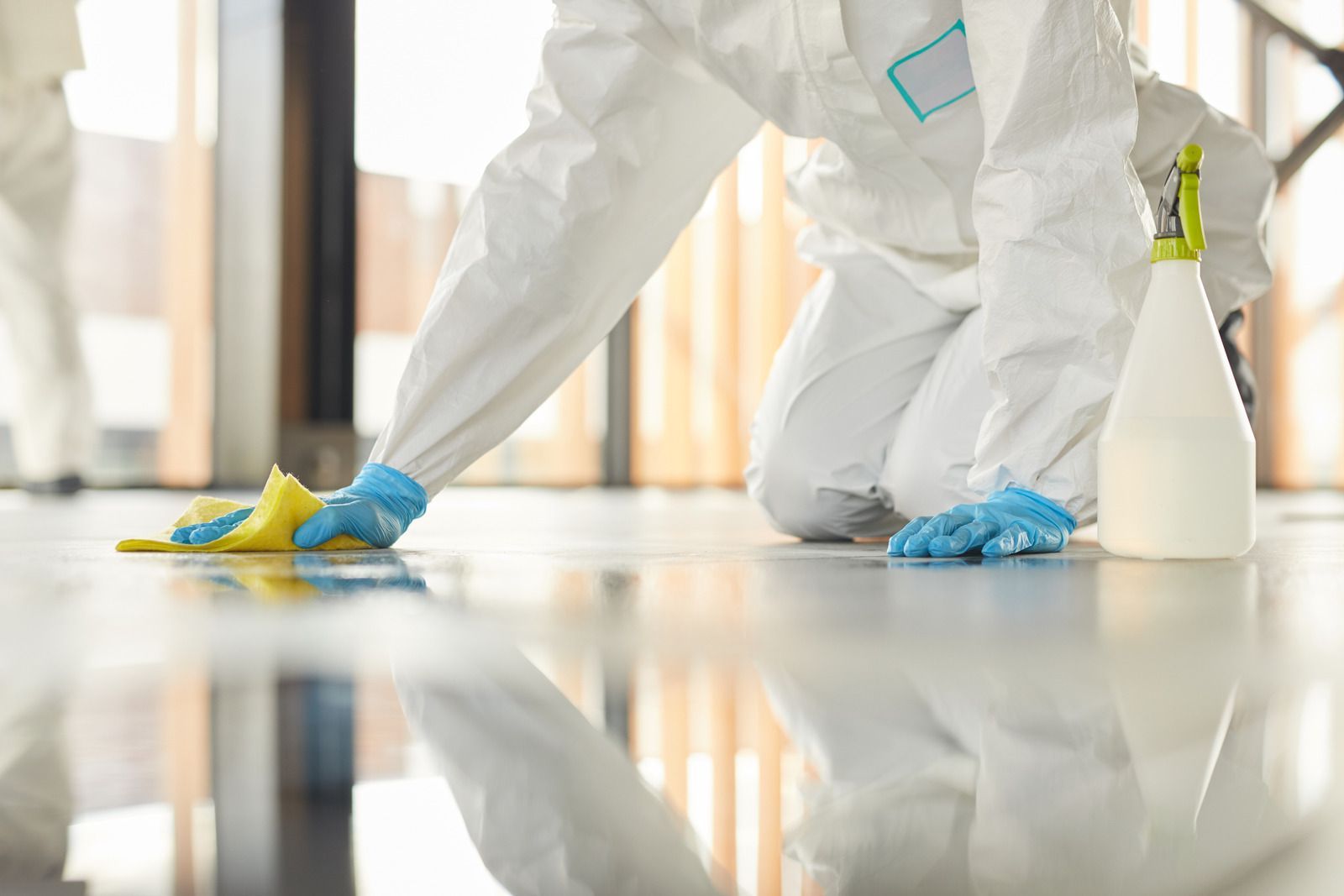 Person in white protective suit kneeling, cleaning floor with sponge and spray bottle.