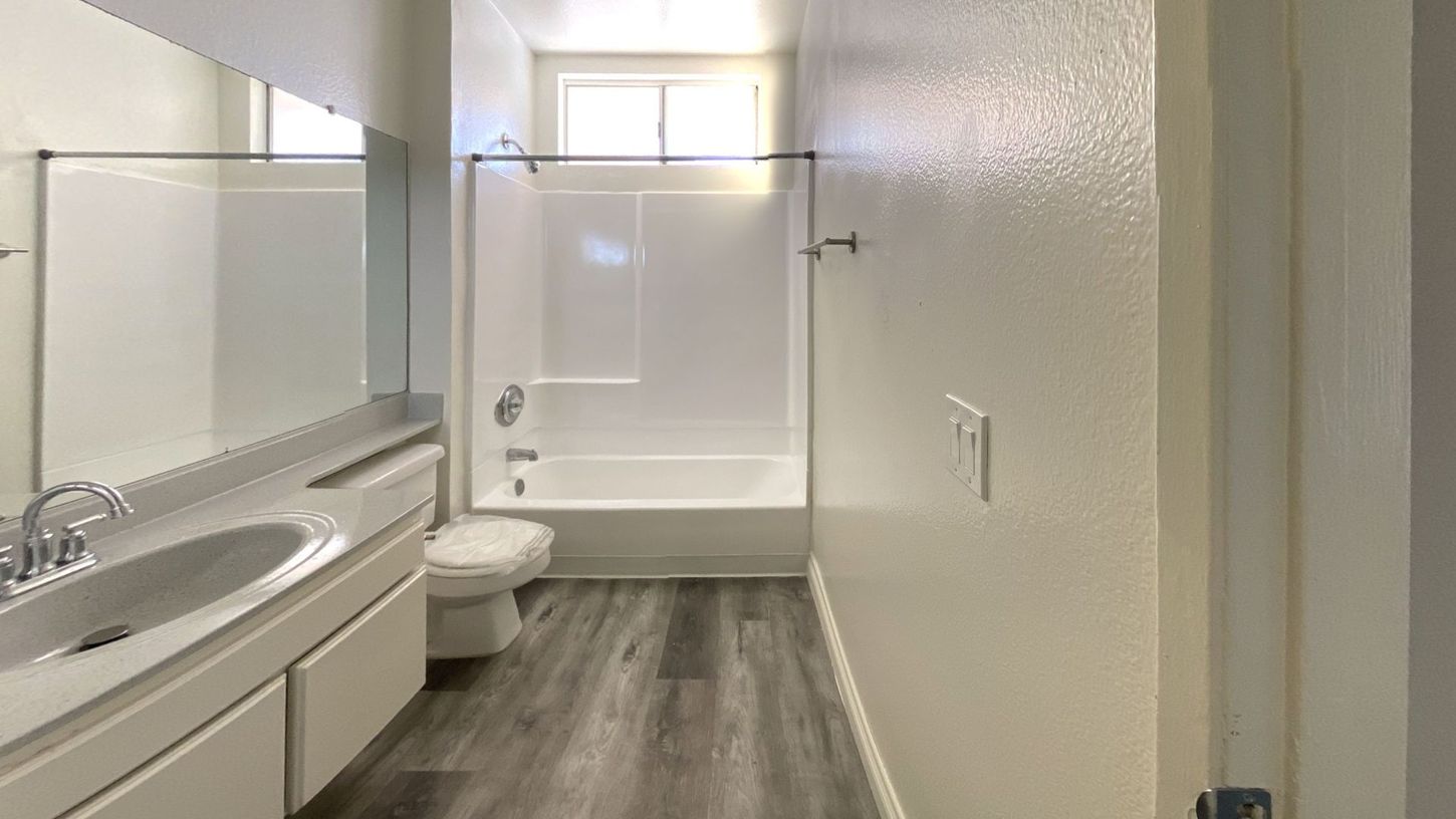 Bathroom with white fixtures, vanity, toilet, and bathtub/shower. Gray wood-look flooring and light-colored walls.