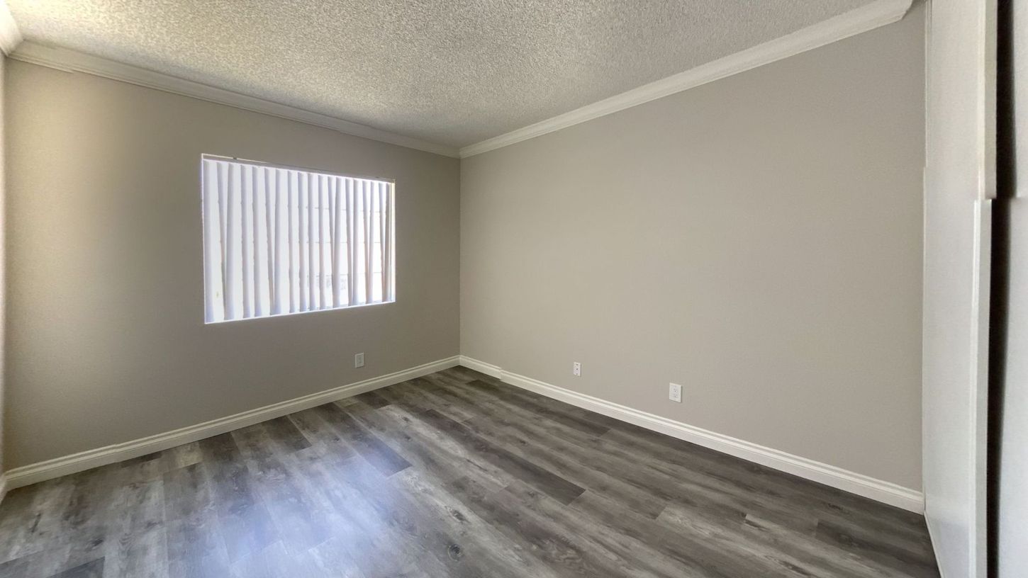 Empty room with wood-look flooring, light gray walls, window with blinds, and white trim.