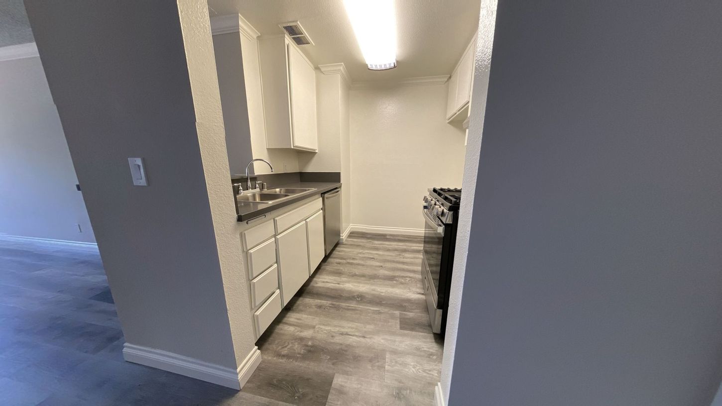 Small, narrow kitchen with white cabinets, gray countertops, and wood-look flooring. A stove is at the end.