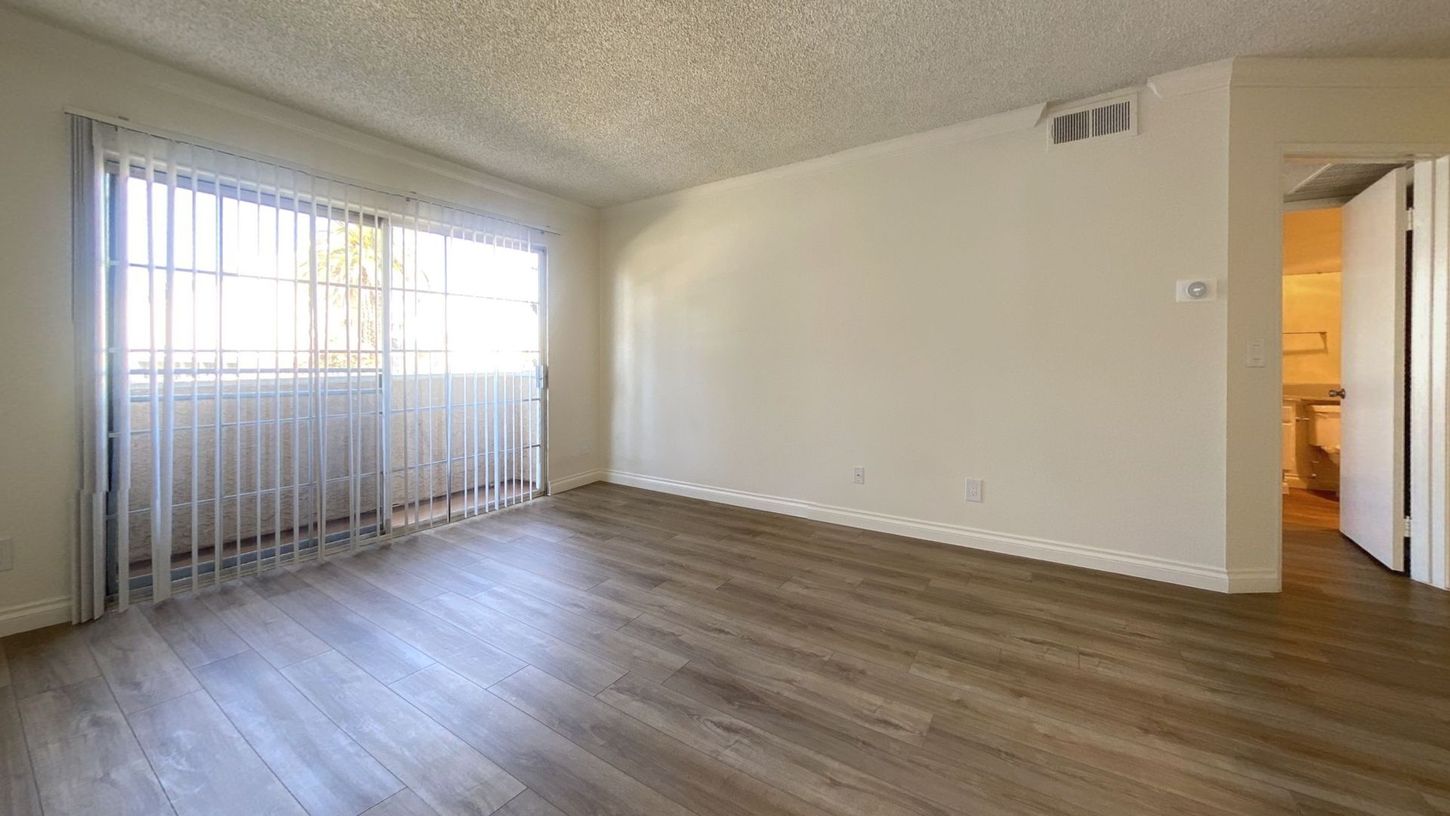 Empty living room with wood-look flooring, large window with blinds, and doorway to a bathroom.