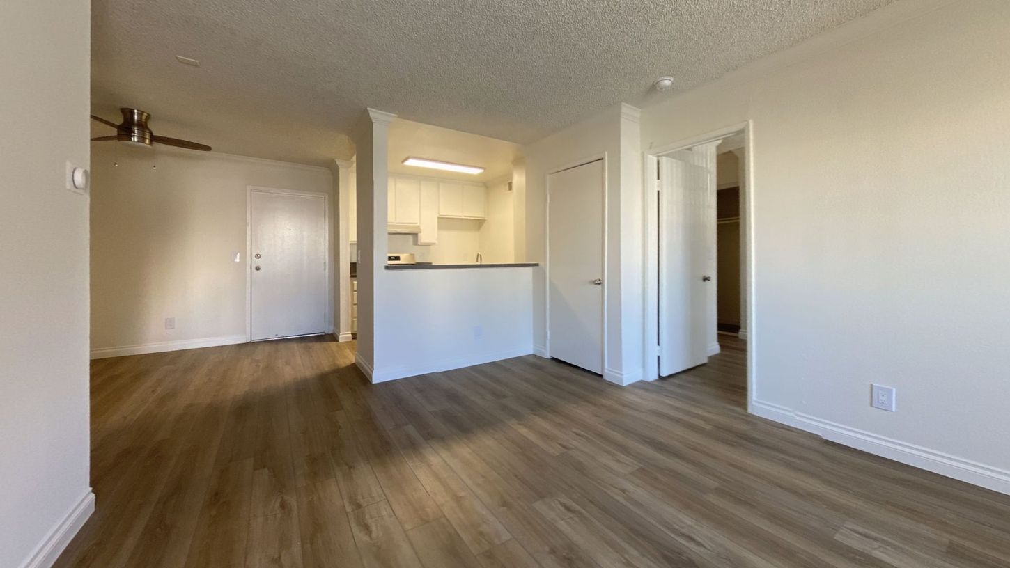 Interior view of an apartment with light wood floors, white walls, and a small kitchen visible in the background.