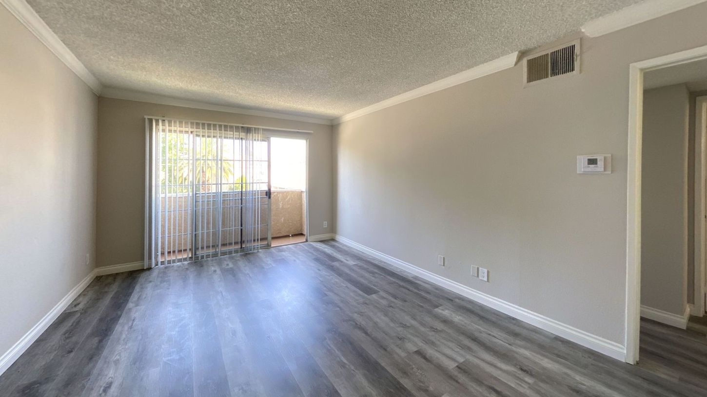 Empty living room with gray wood-look flooring, light gray walls, and sliding glass door to a balcony.
