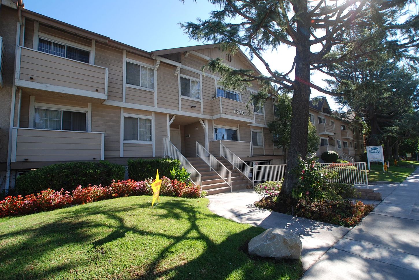 Apartment building exterior, beige siding, stair access, tree shadows on lawn, sunny day.