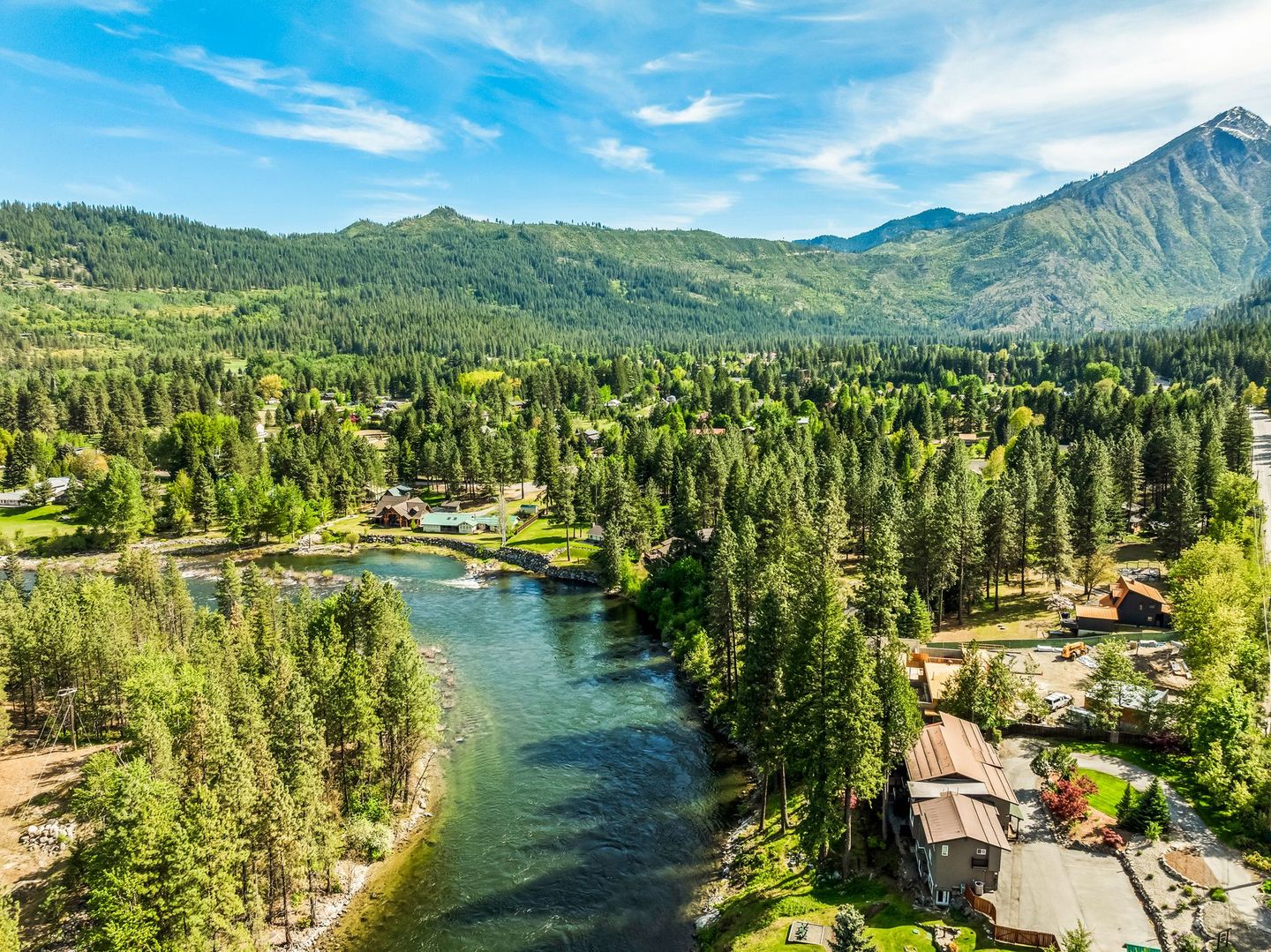 River winding through a lush green valley with evergreen trees, buildings, and mountains under a blue sky.