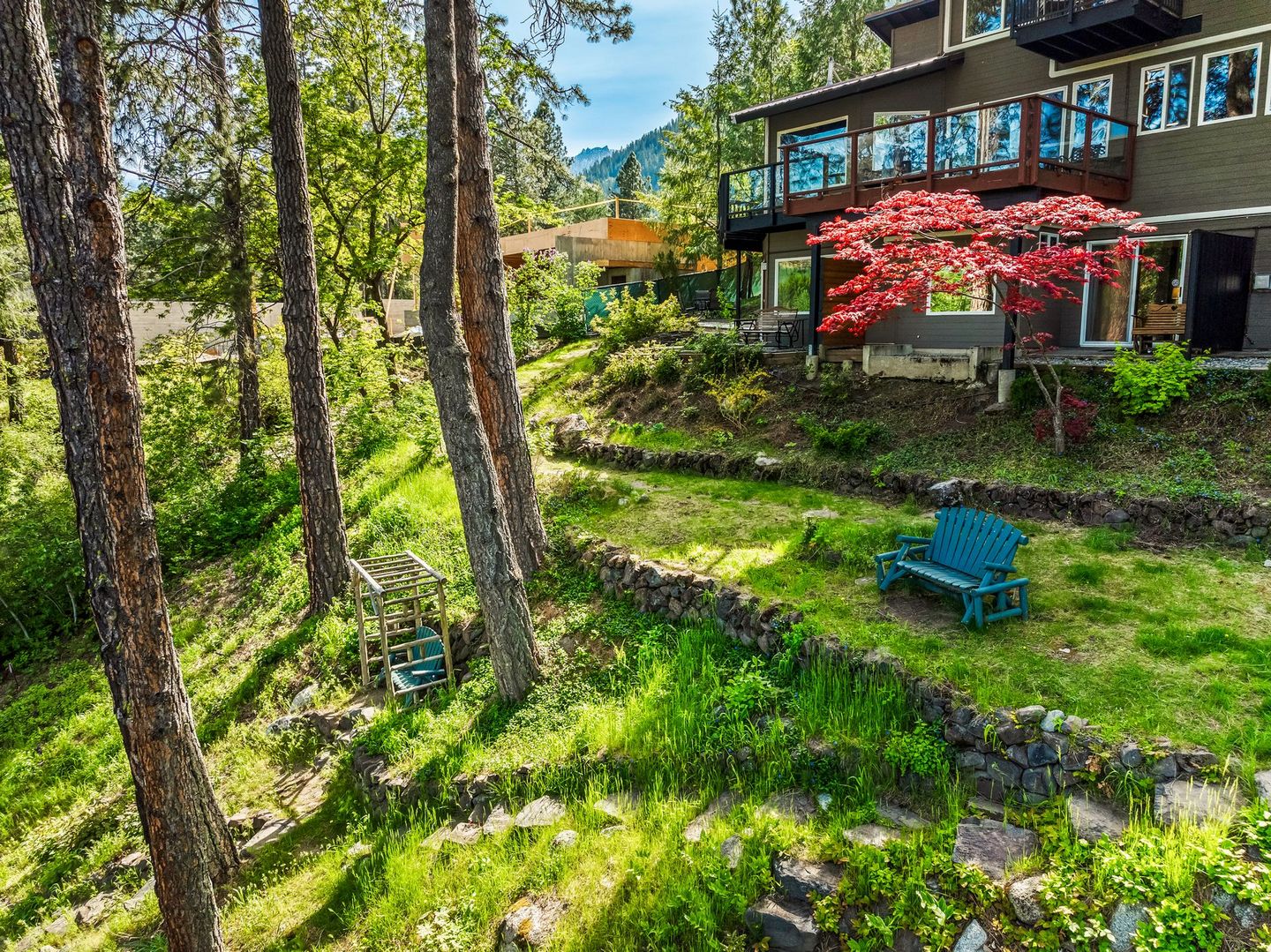 Hotel with multiple levels, balcony, and red-leafed tree. Green grass, stone walls, and a blue bench are visible.