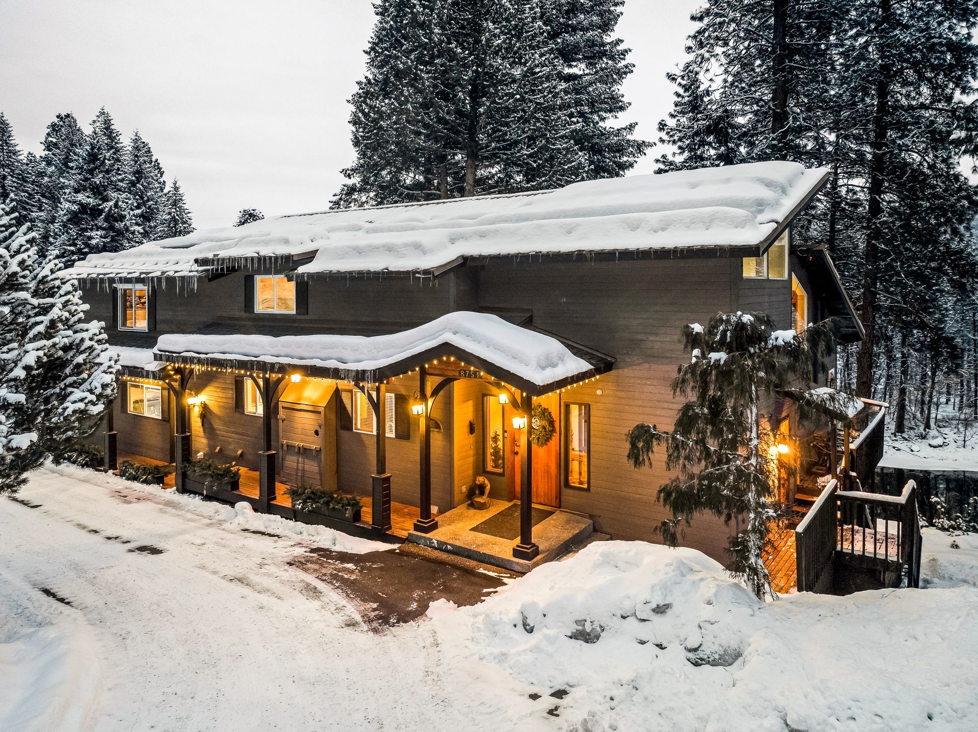 Snow-covered house with porch lights on; surrounded by snow-covered trees and ground.