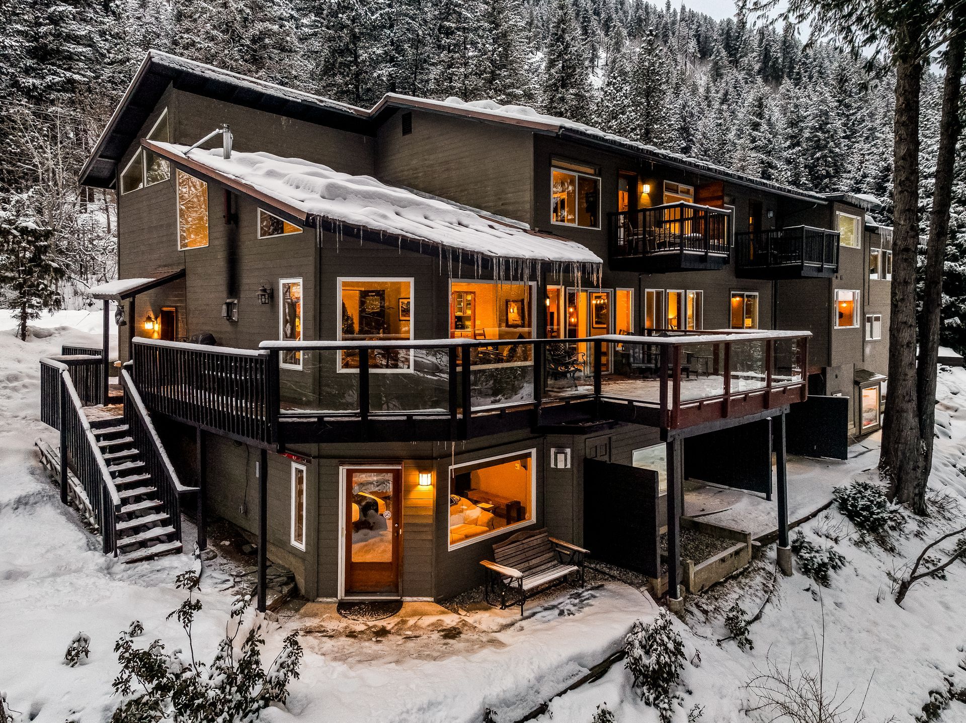 Two-story house with snow-covered roof and deck, surrounded by snow and trees; lights on inside.
