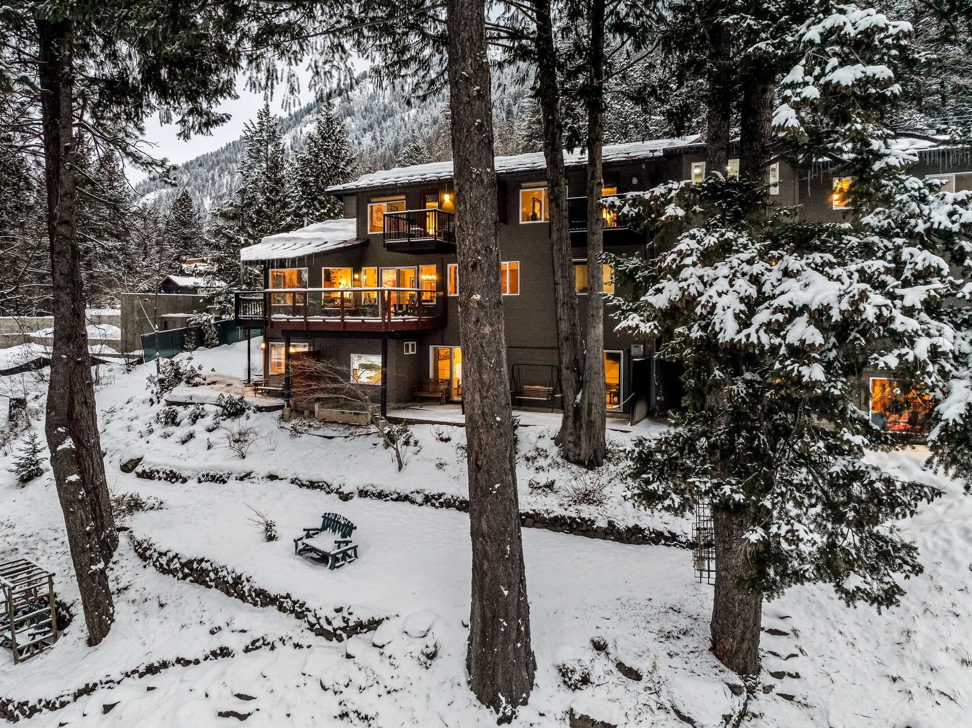 Snow-covered house nestled among trees, windows lit, a fire pit in the yard.