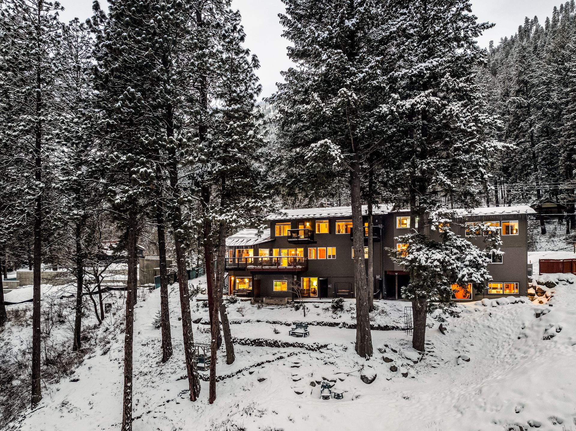 Snow-covered home nestled among tall pine trees on a hillside; lights glowing inside, winter setting.