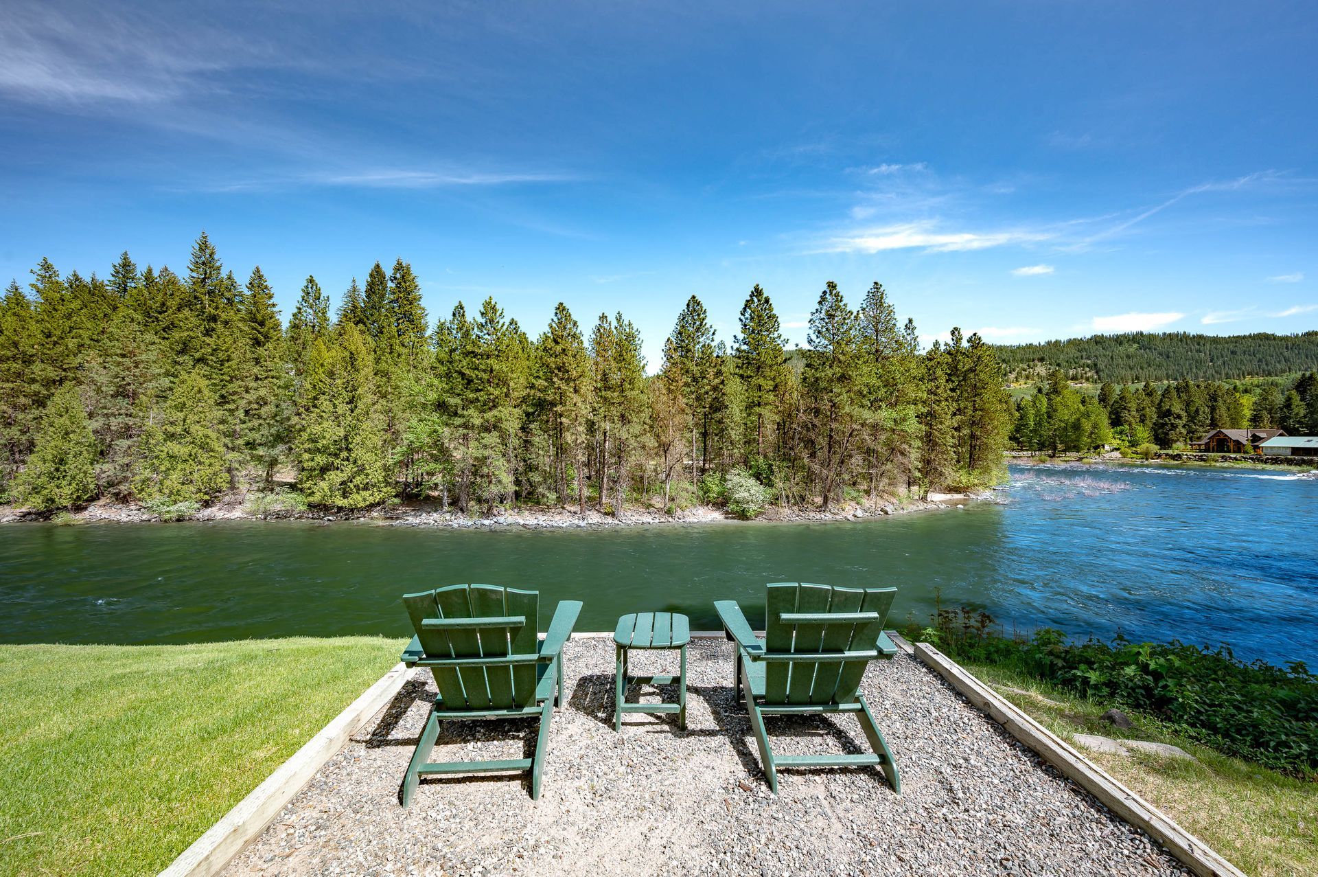 Two green Adirondack chairs and small table on gravel overlooking a lake and forested shoreline.