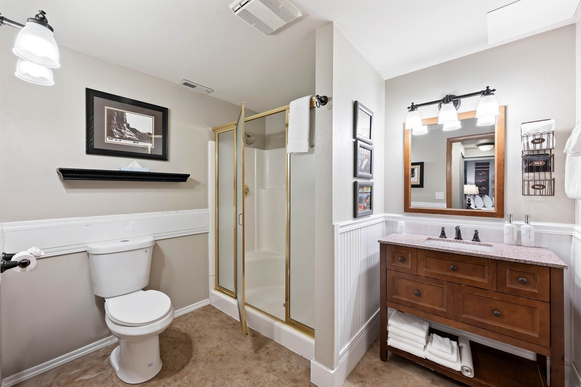 Bathroom with toilet, shower, wooden vanity, framed art, and white paneling.