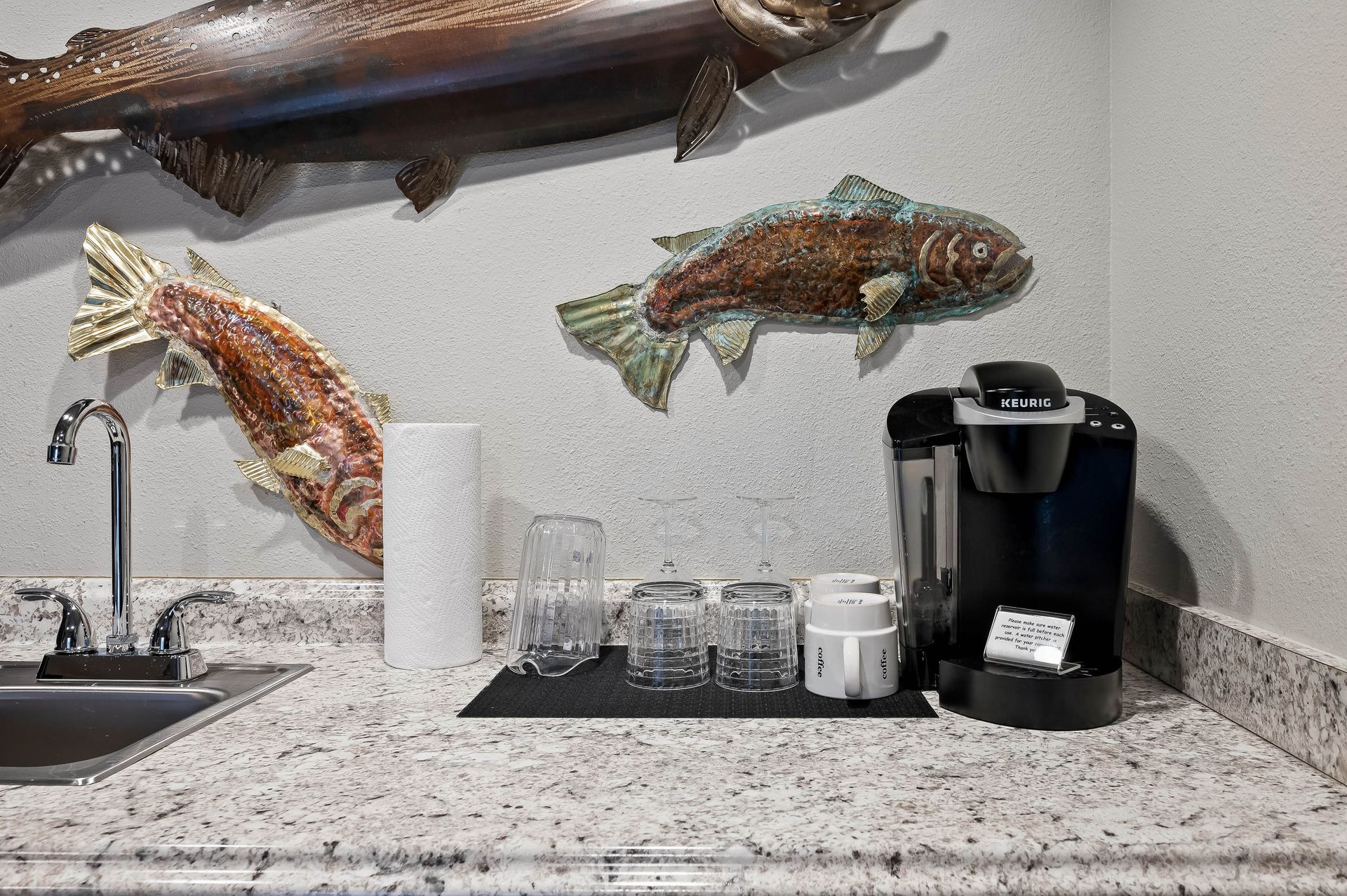 Countertop with sink, coffee maker, glasses, and fish art on wall.