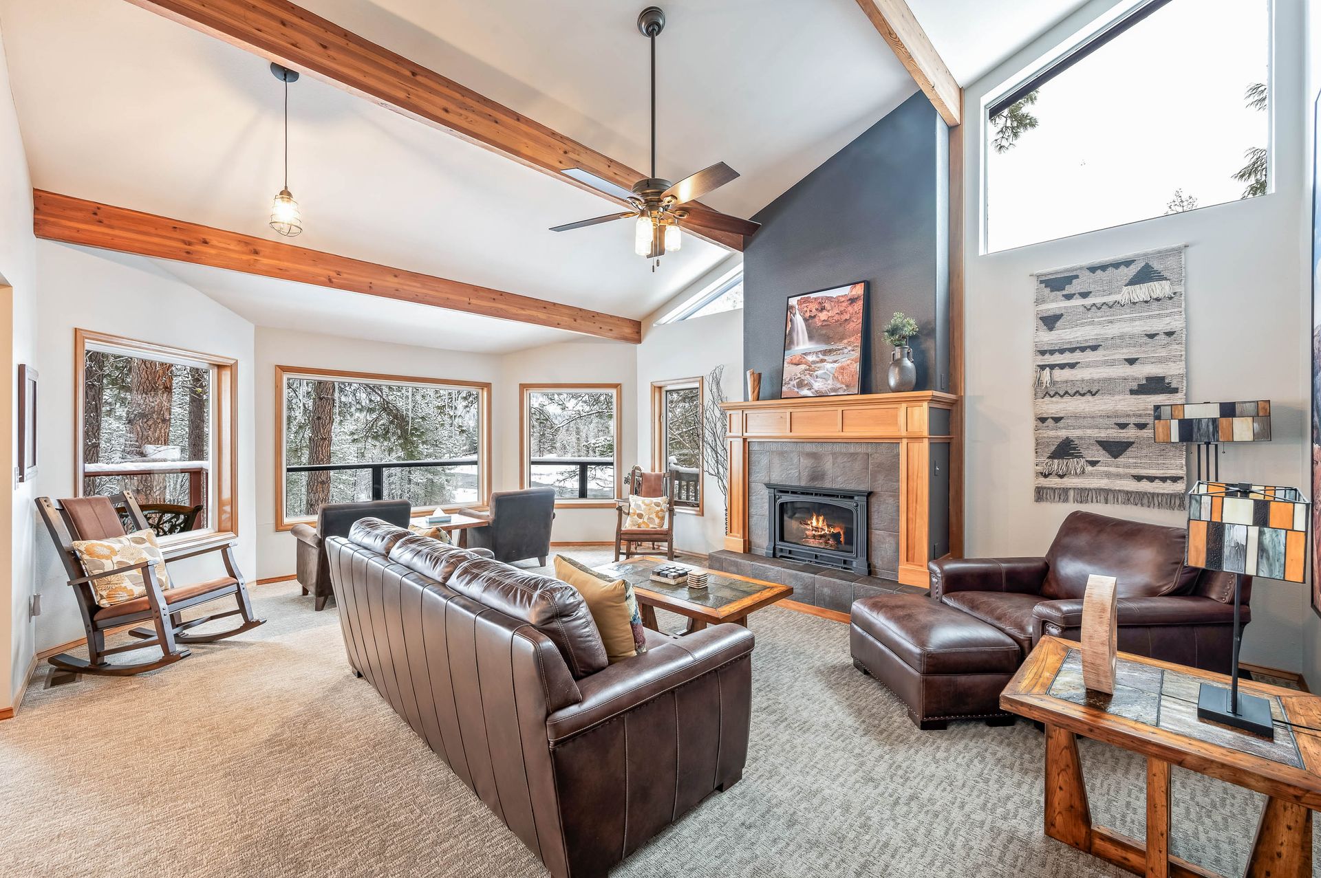 Living room with brown leather sofa, fireplace, and large windows with a wooded view.