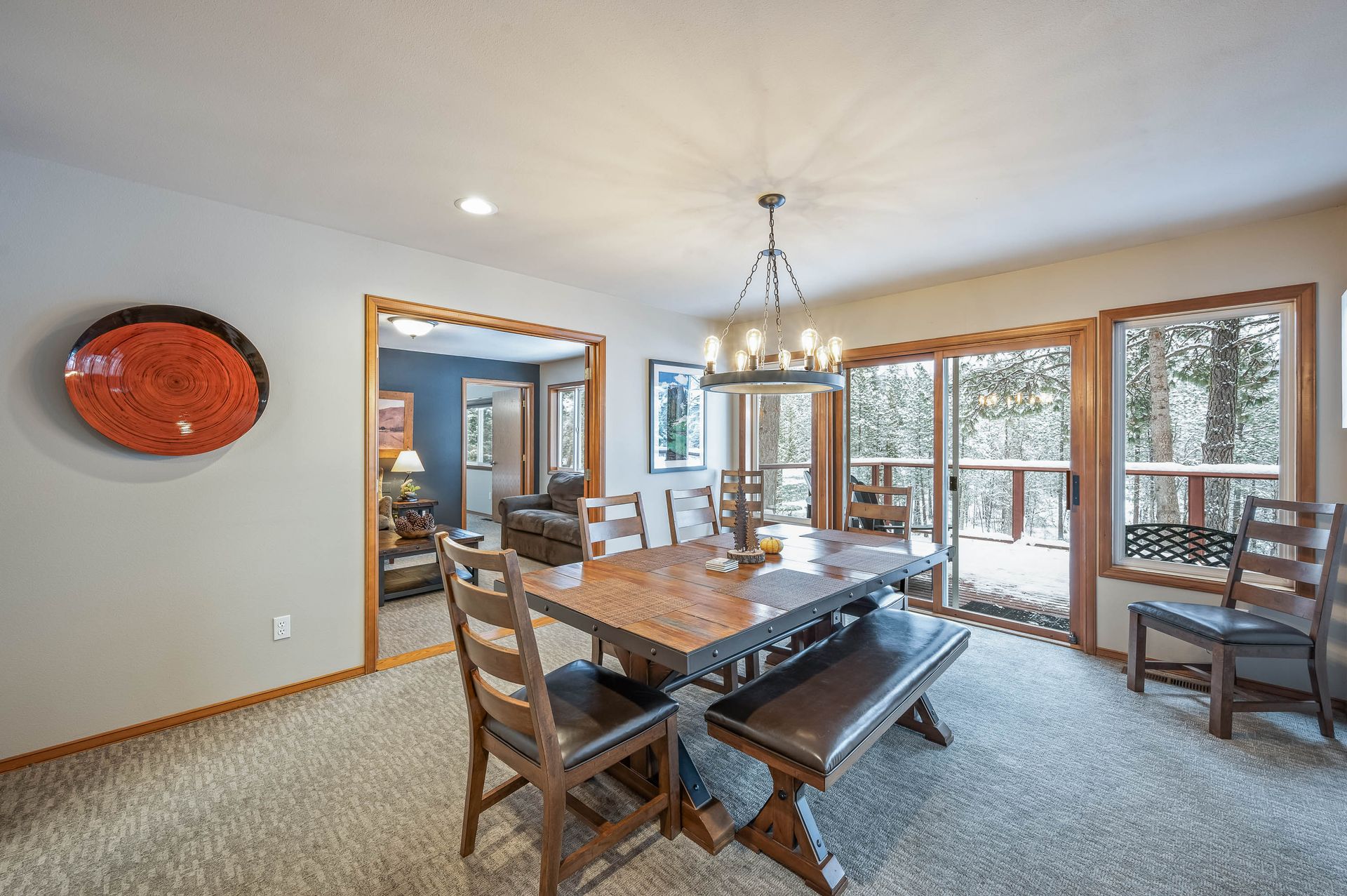 Dining room with wooden table, bench, chairs, and large windows overlooking a deck and trees.