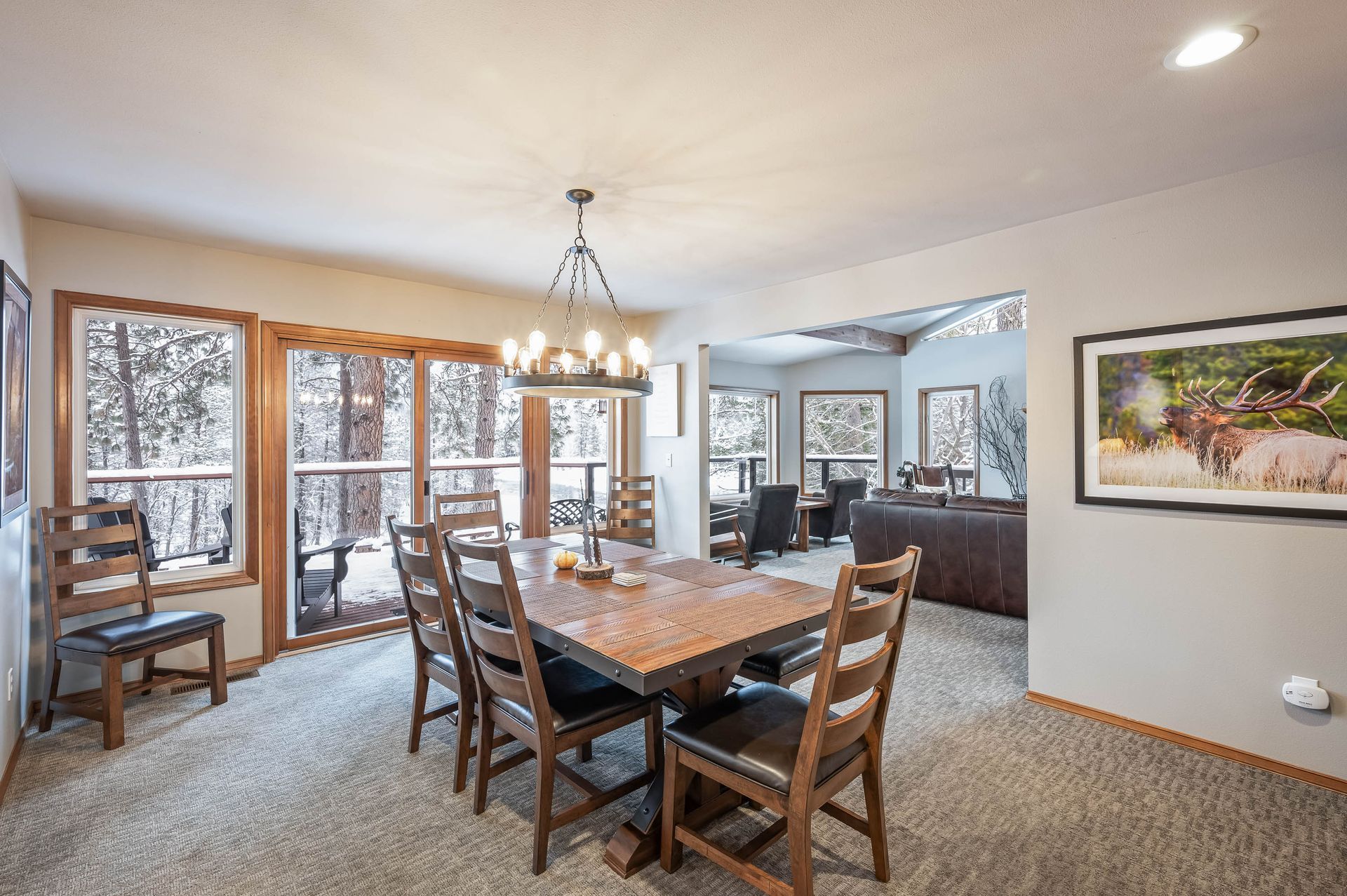 Dining room with wooden table and chairs, large windows with snowy trees, and open to a living area.