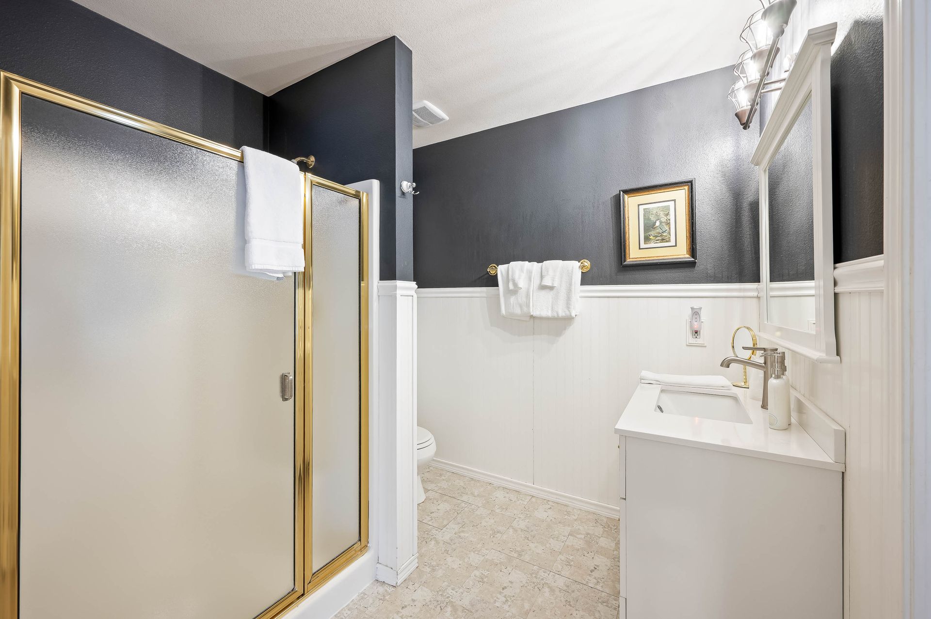 Bathroom with frosted glass shower, gold trim, white vanity, dark blue wall, and painting.