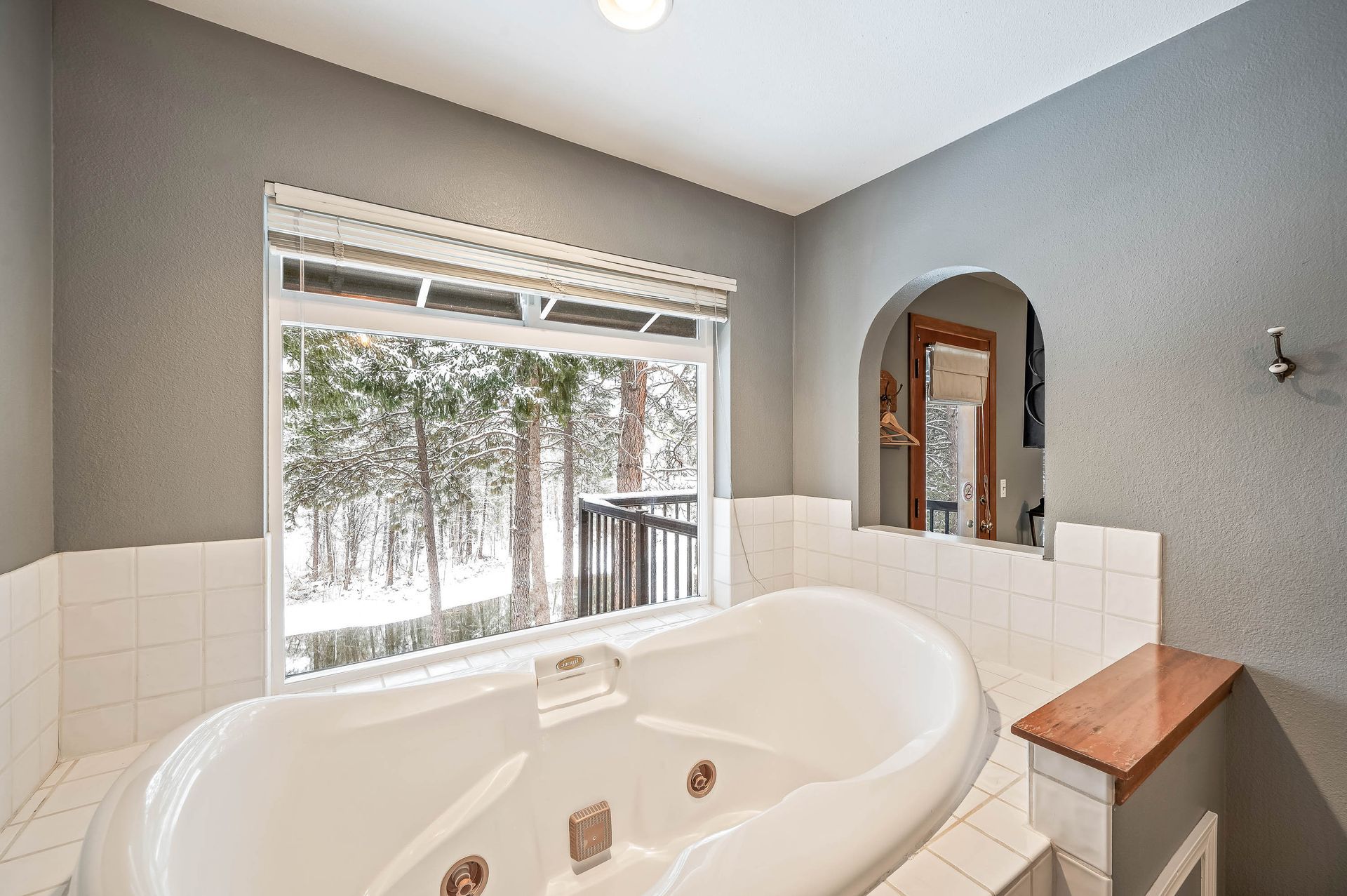 Bathroom with a white jacuzzi tub, window overlooking snowy trees, gray walls, and an arched alcove with mirror.