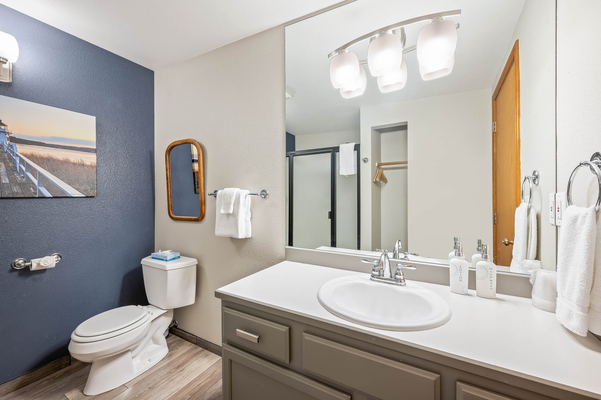 Bathroom with blue accent wall, white vanity, large mirror, toilet, shower, and artwork.