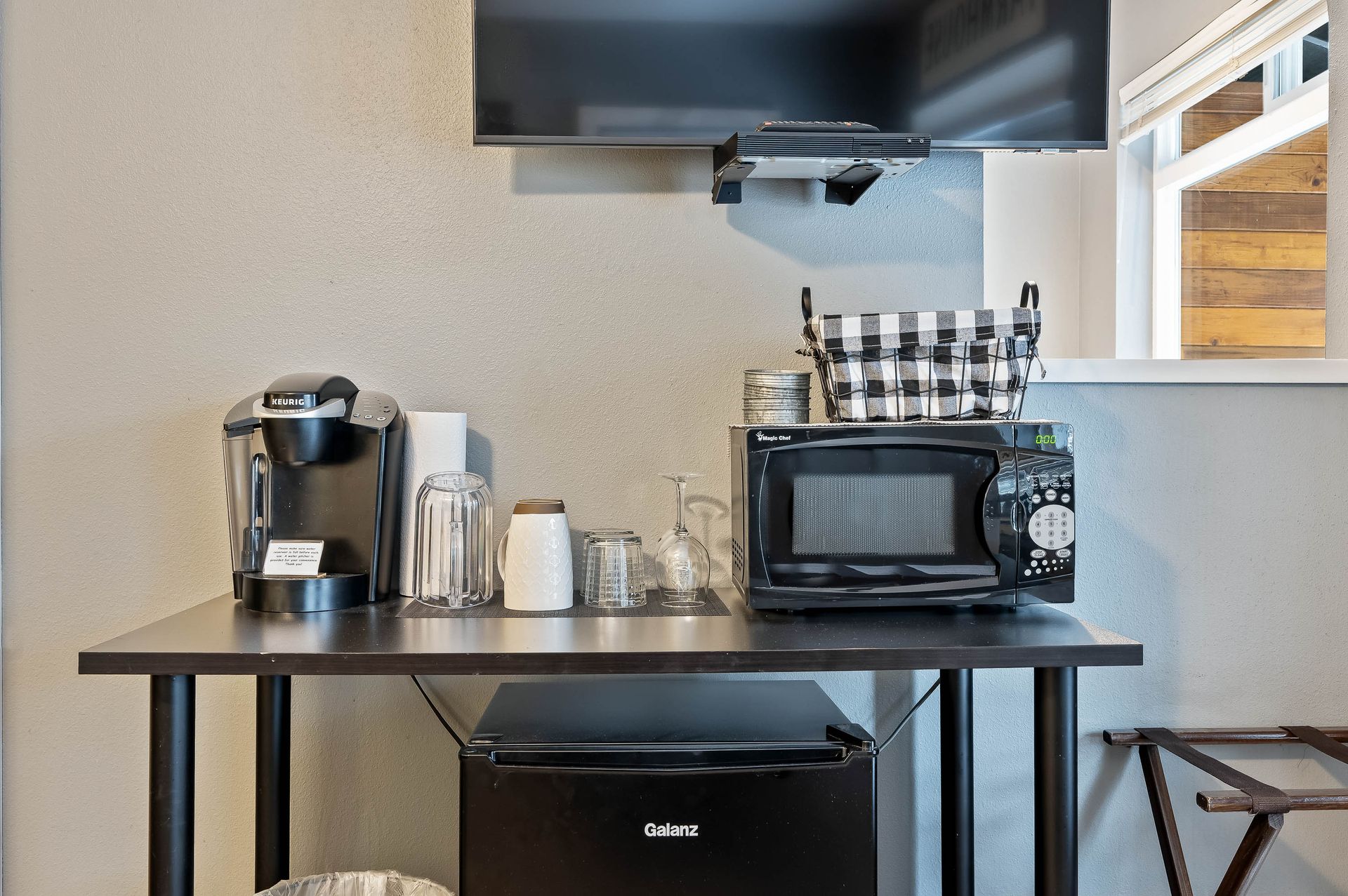 Black table with coffee maker, microwave, mini-fridge, glasses, TV above, and window.