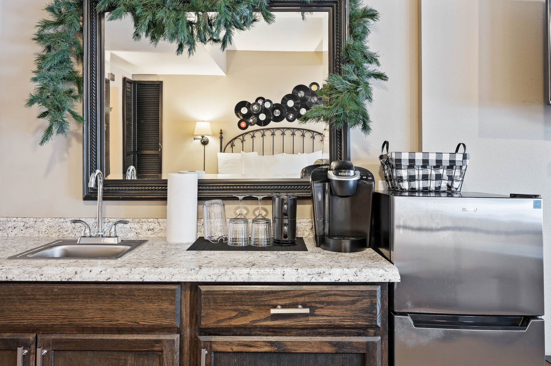 Countertop with sink, Keurig, glasses, fridge, and mirror decorated with greenery.