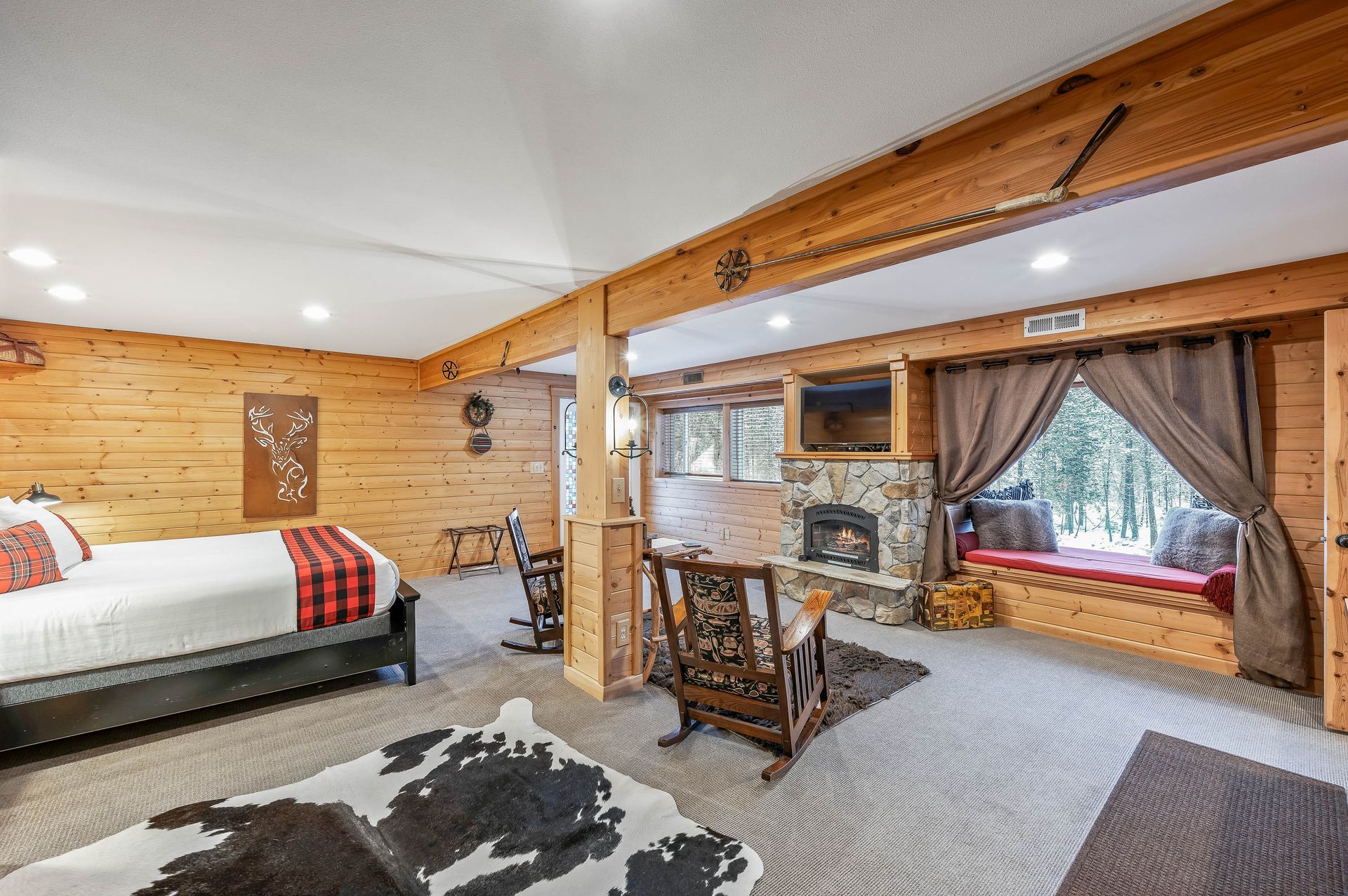 Cozy cabin bedroom with a bed, fireplace, and window seat overlooking a snowy forest.