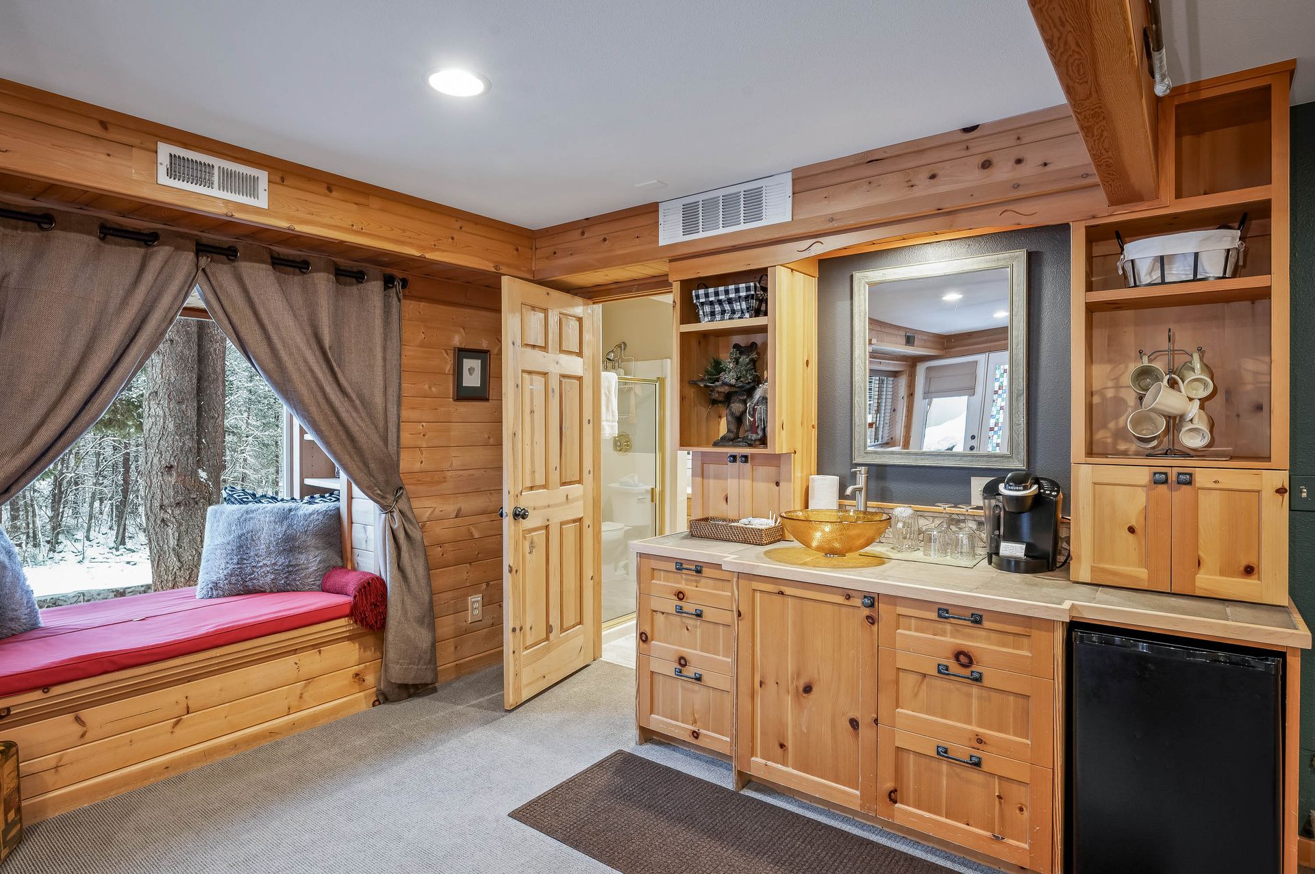 Cozy wooden room with built-in cabinetry, window seat, and small fridge. Light wood paneling, mirror, and coffee station.