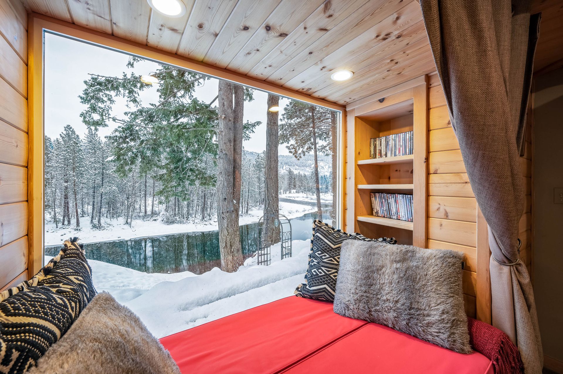 Cozy cabin interior with a large window overlooking a snowy forest. Red seating, pillows, and bookshelf.