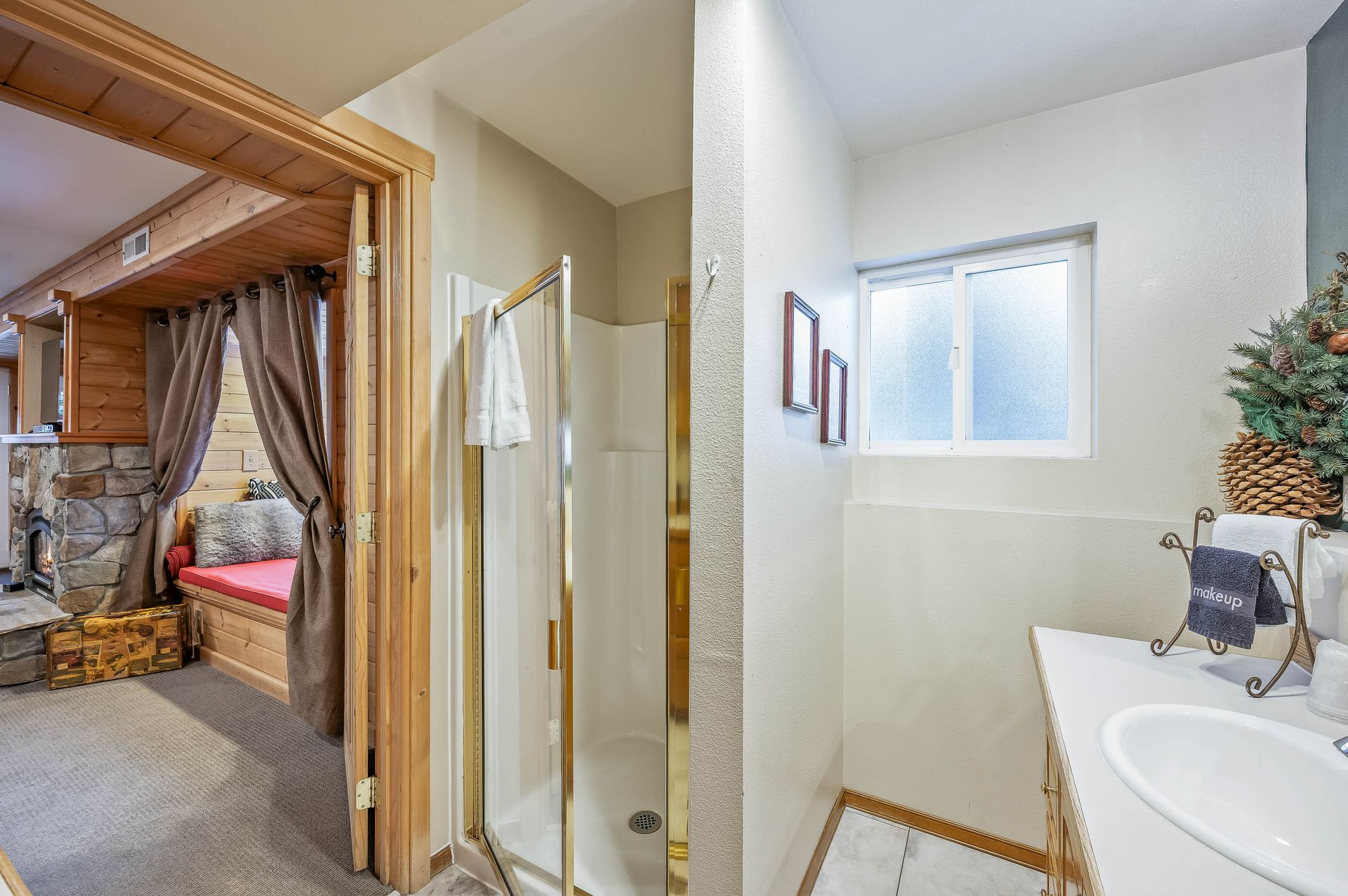 Bathroom with shower, vanity, and window. Another room with fireplace, bench, and curtains is visible through the doorway.