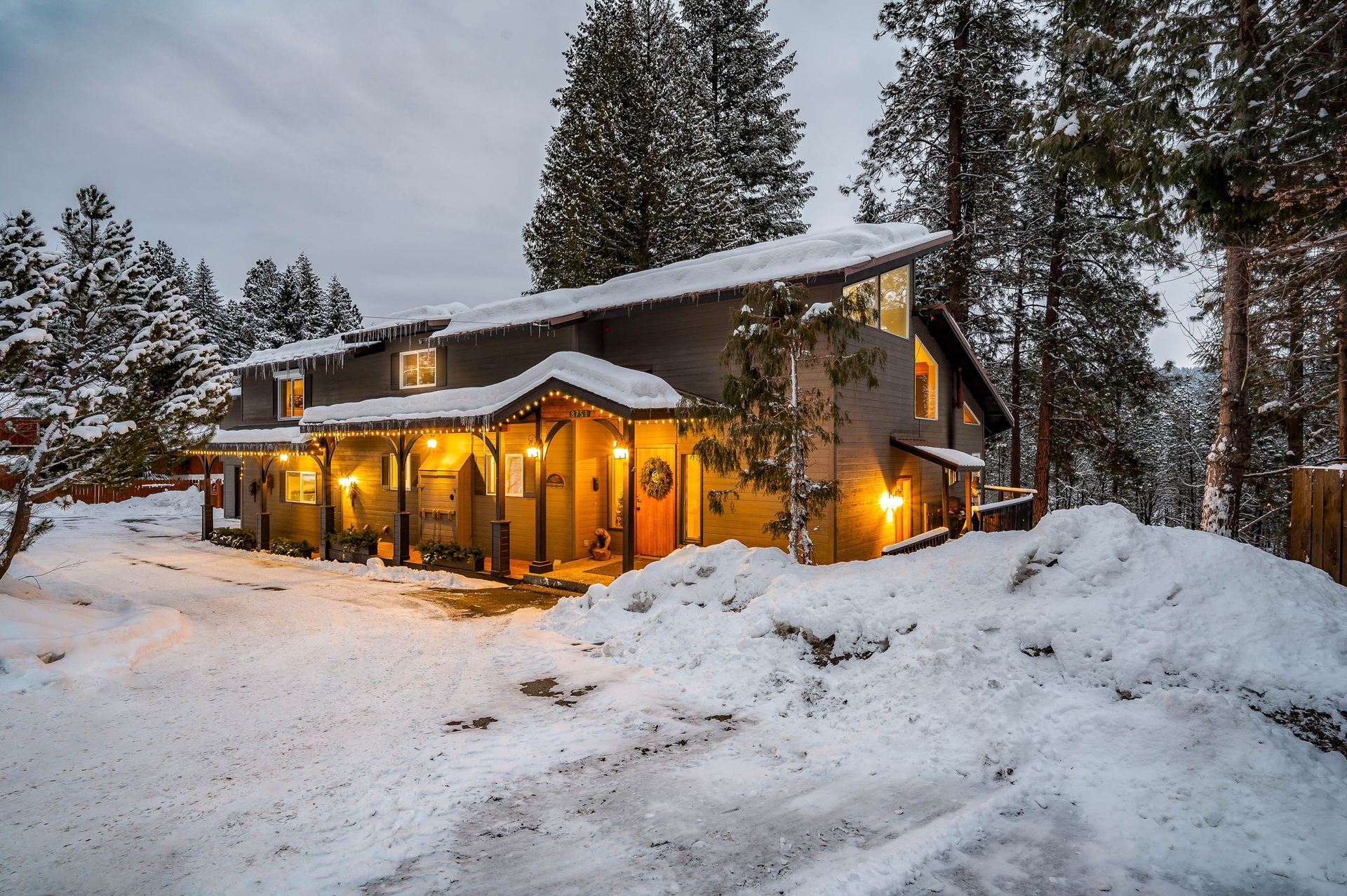 Snowy house with sloped roof, covered in snow, surrounded by pine trees, lit by warm lights.