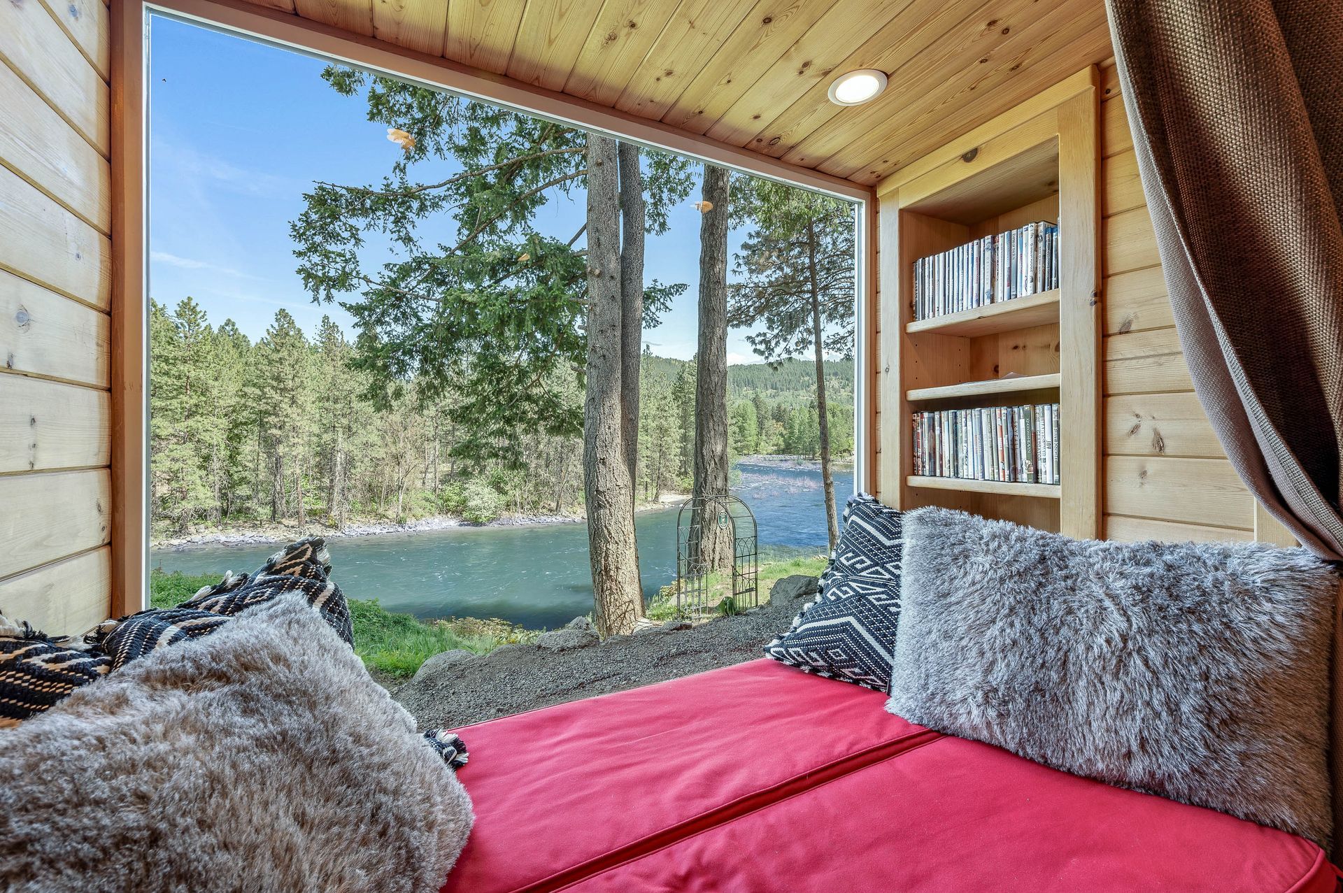 Cozy nook with red cushion, fluffy pillows, bookshelf, and large window overlooking a river and trees.