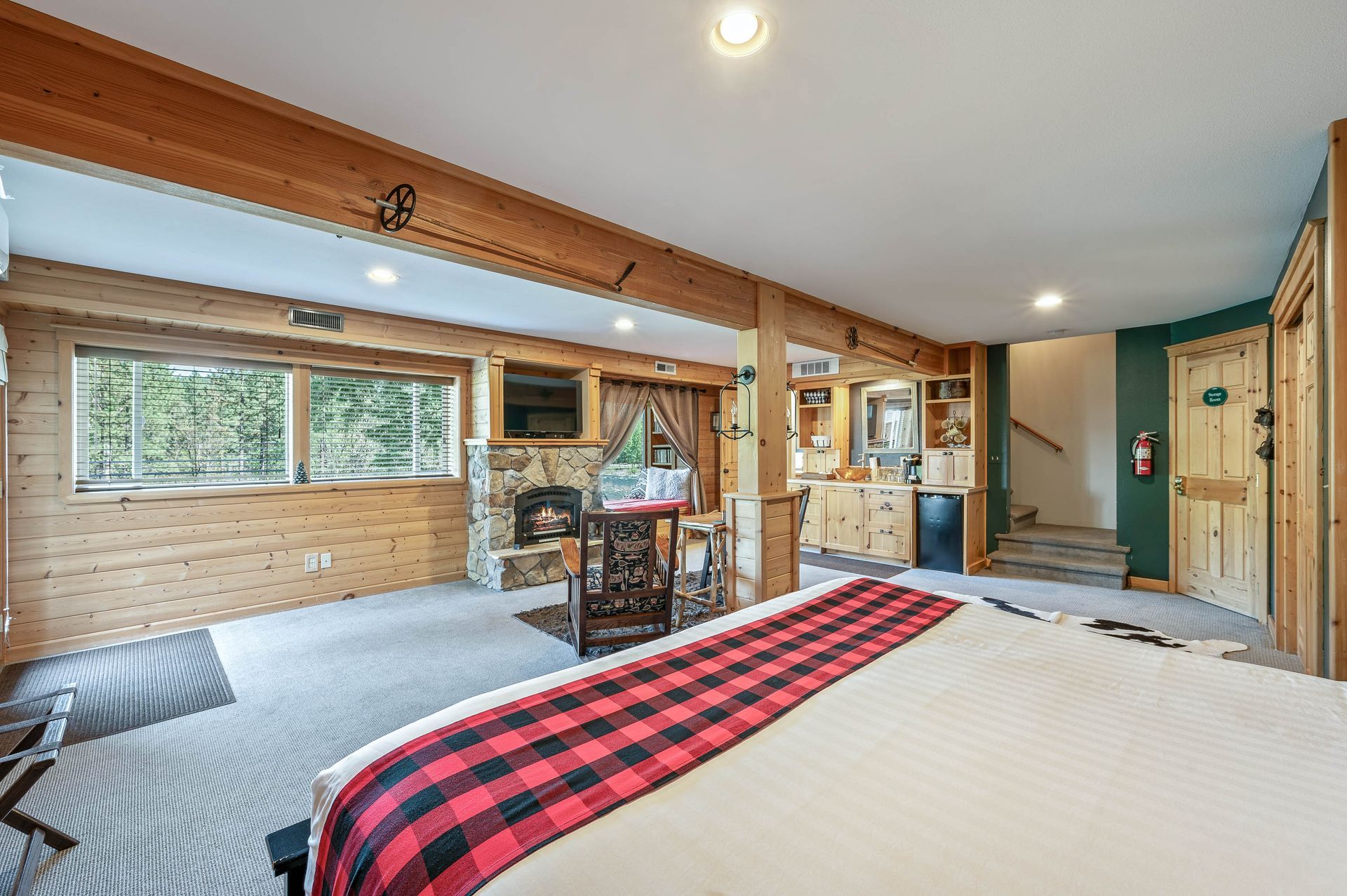 Cozy cabin bedroom with wood beams, fireplace, bed with red plaid blanket, and small table.