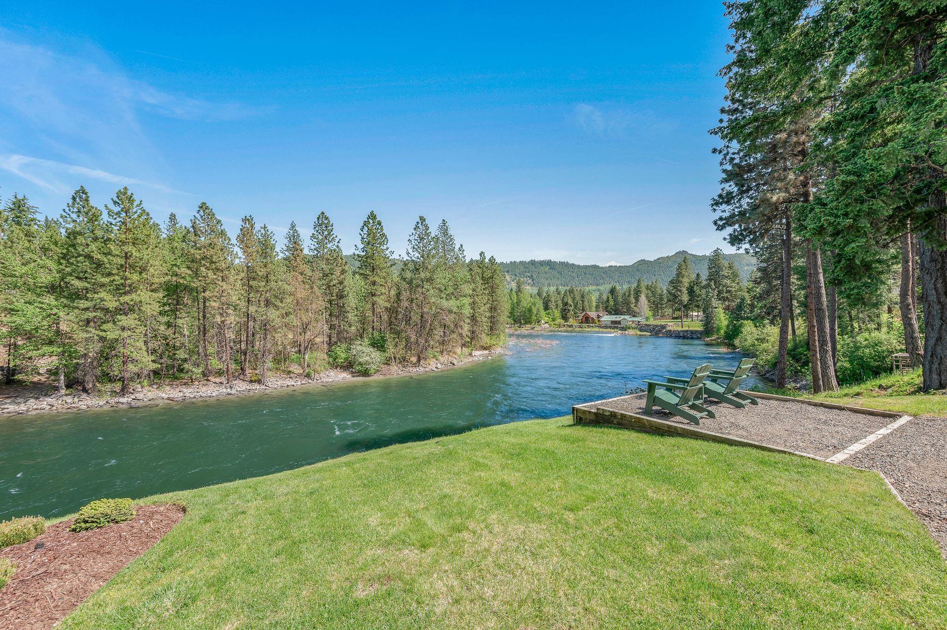 A river flows through a forest under a blue sky, with a grassy area in the foreground.