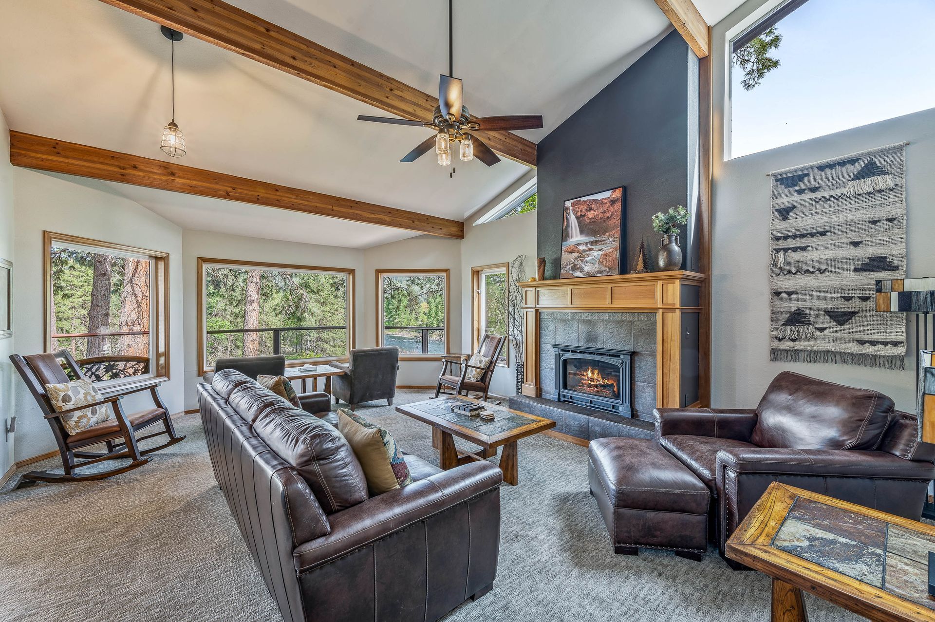 Cozy living room with vaulted ceiling, exposed beams, fireplace, leather furniture, windows, and decorative wall hanging.