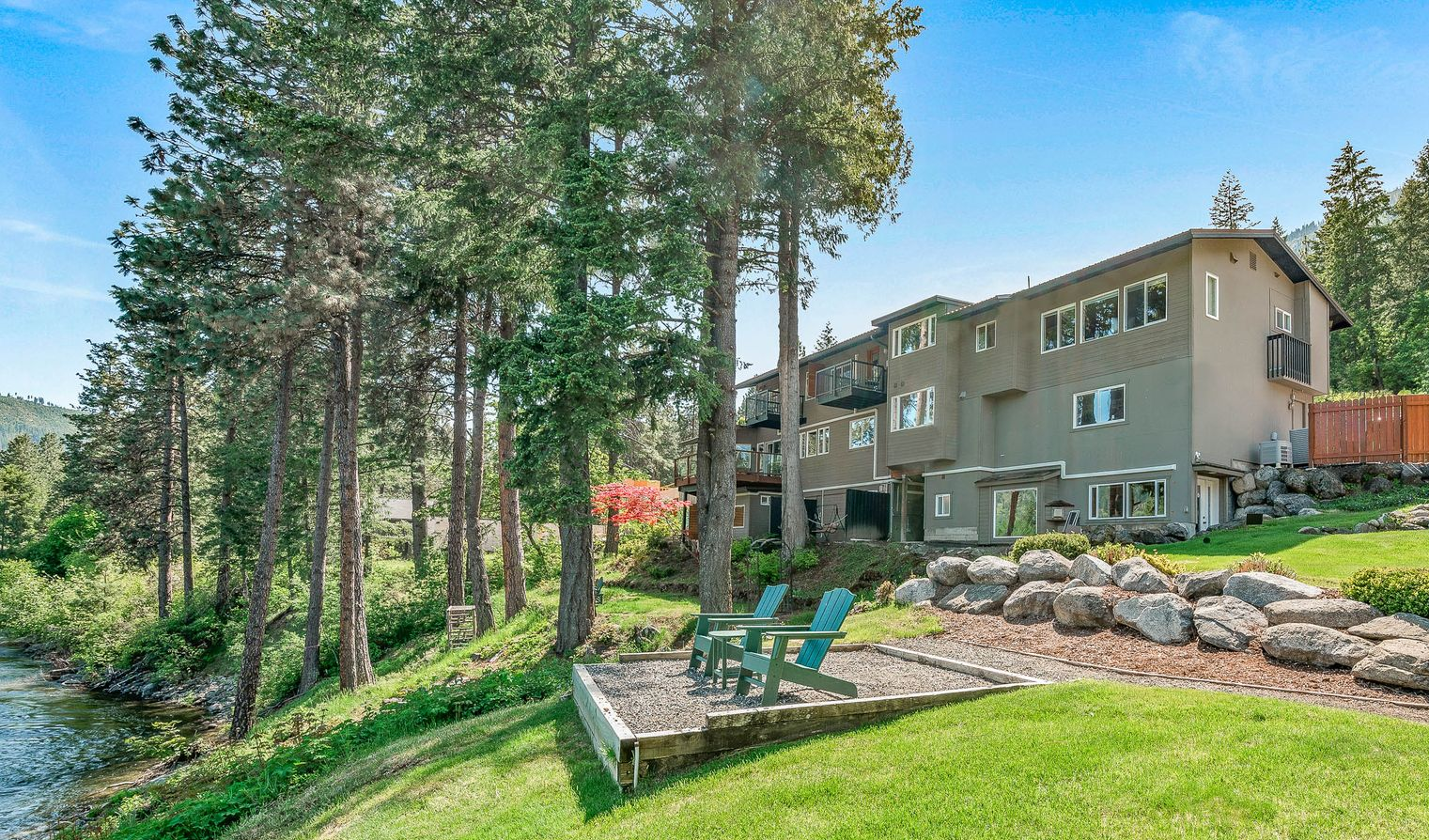 Lakeside apartment building with green lawn, trees, and blue chairs.