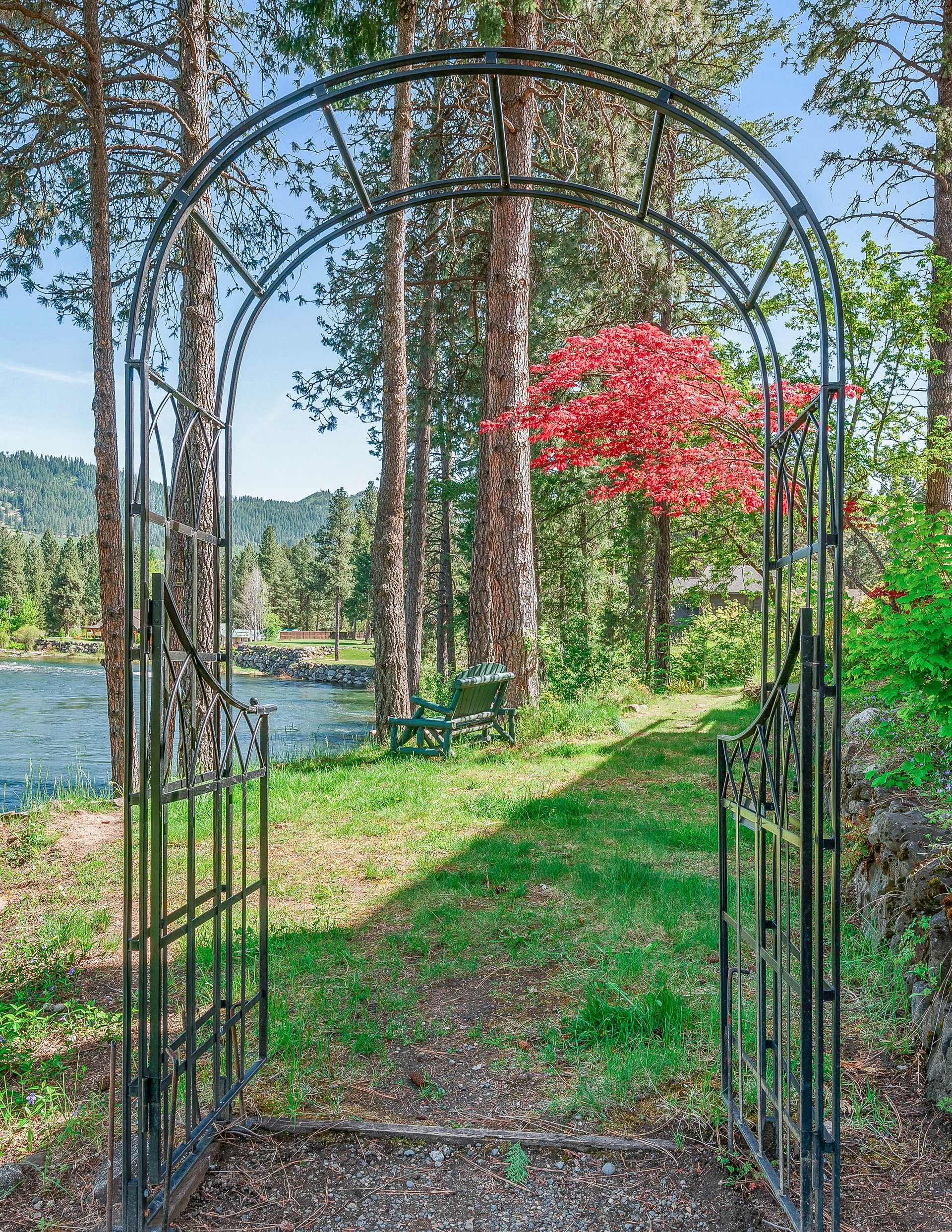 Black metal garden archway overlooking a river, with red tree and forest background.