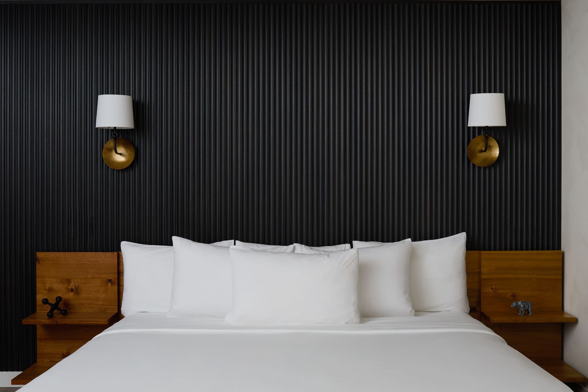 Modern bed with white linens against a dark striped wall, flanked by wooden nightstands and wall sconces
