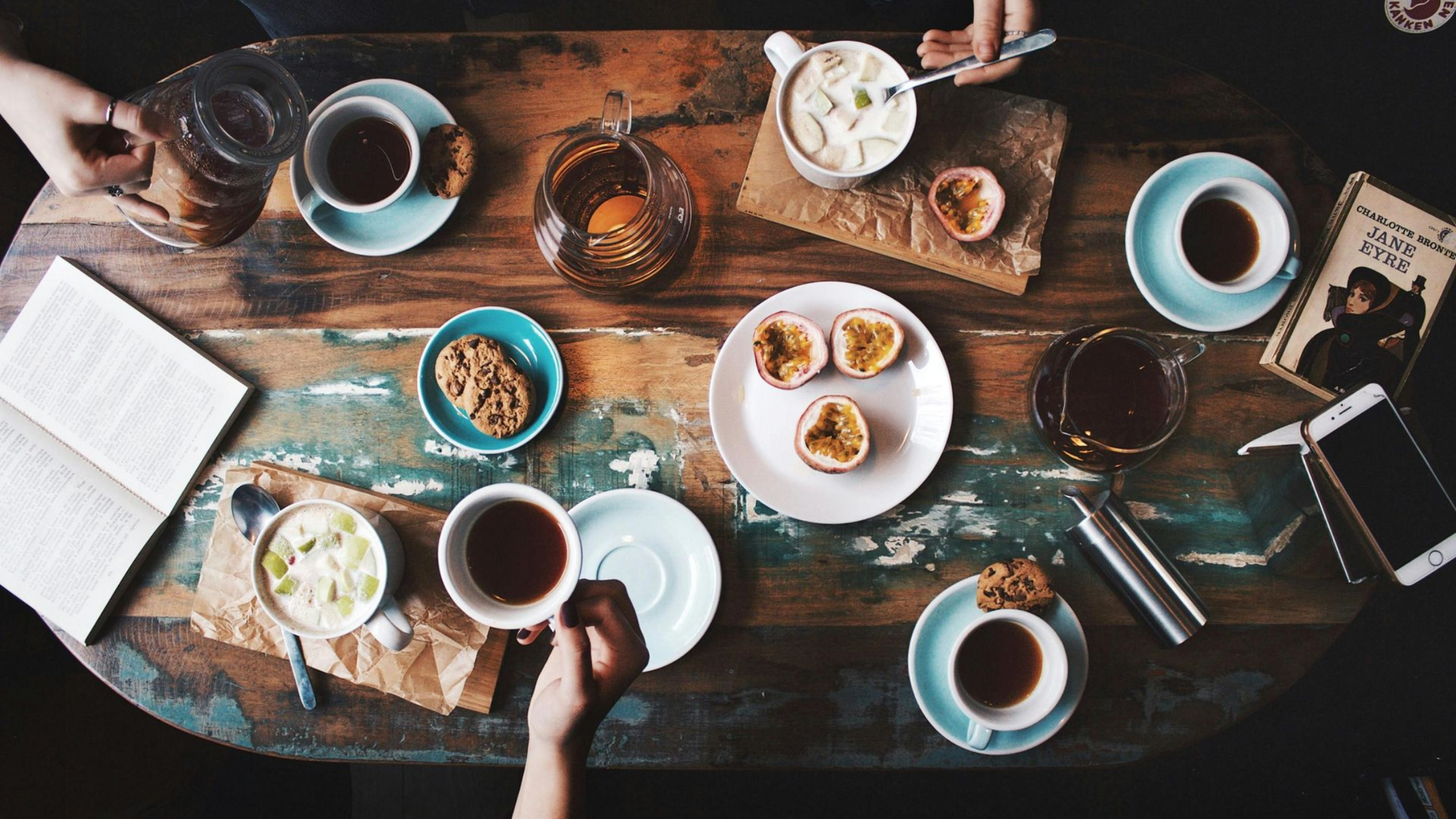Overhead view of a rustic table with drinks, food (passionfruit, oatmeal), book, phone, and two people’s hands.