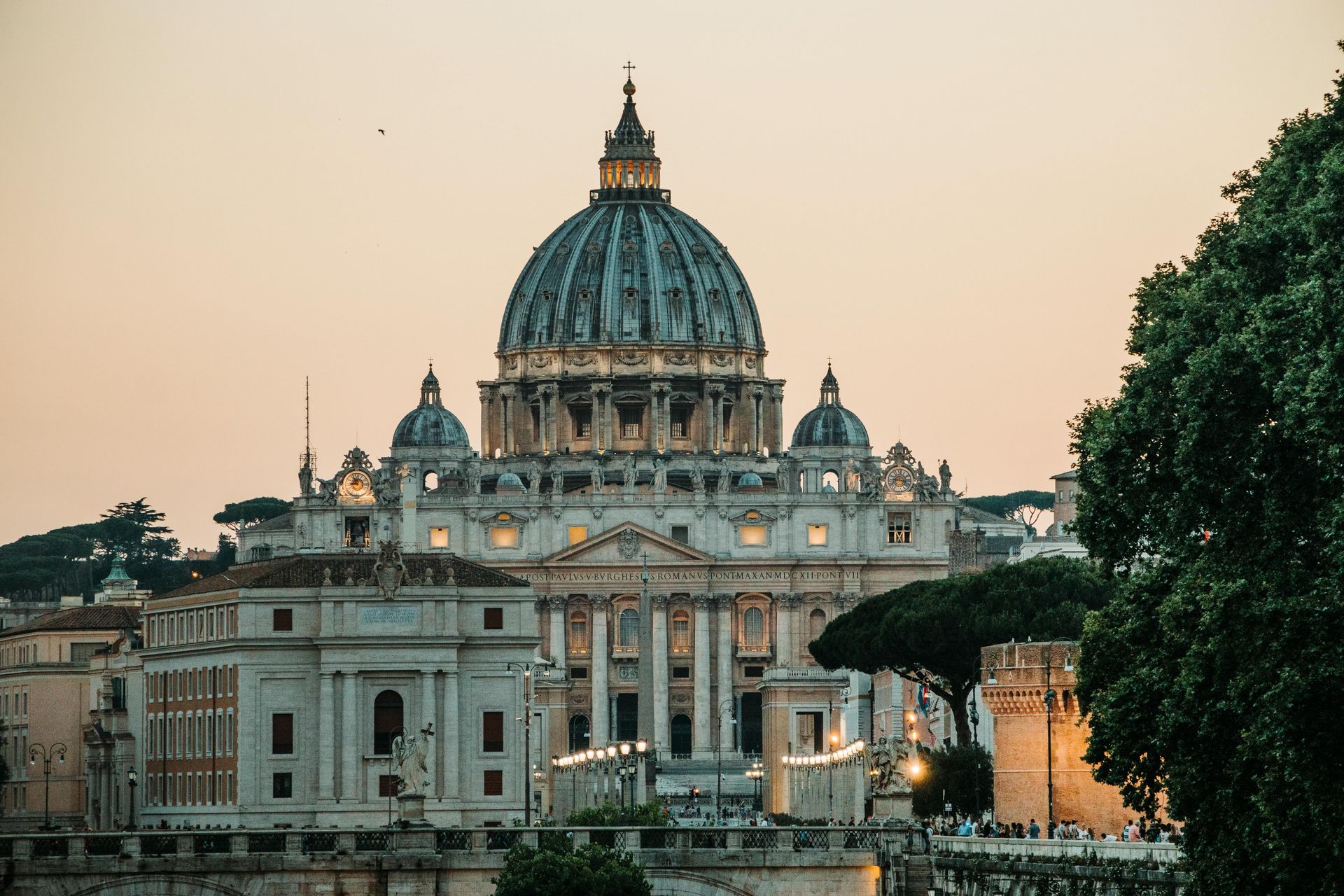 A view of St. Peter's Basilica at sunset, with trees and historical buildings surrounding the church.