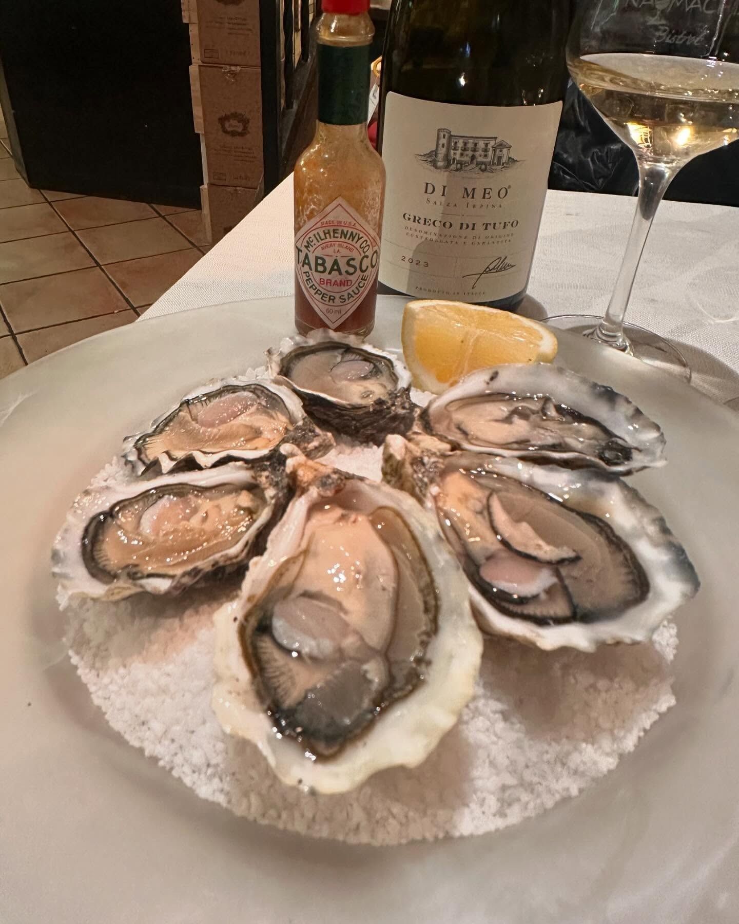 Un piatto di ostriche su ghiaccio con spicchi di limone, salsa piccante e un bicchiere di vino bianco su un tavolo.