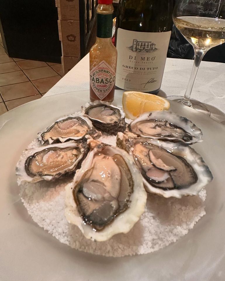 Un piatto di ostriche su ghiaccio con spicchi di limone, salsa piccante e un bicchiere di vino bianco su un tavolo.