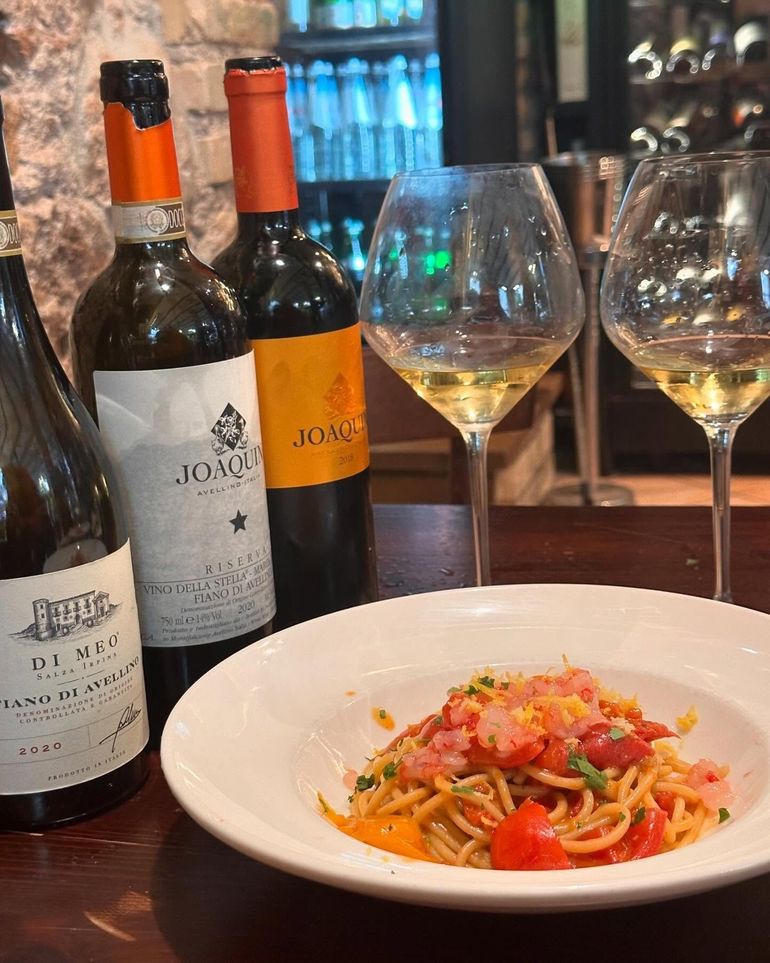 Cena a base di pasta con bottiglie e bicchieri di vino su un tavolo di ristorante, e una ciotola bianca di pasta davanti.