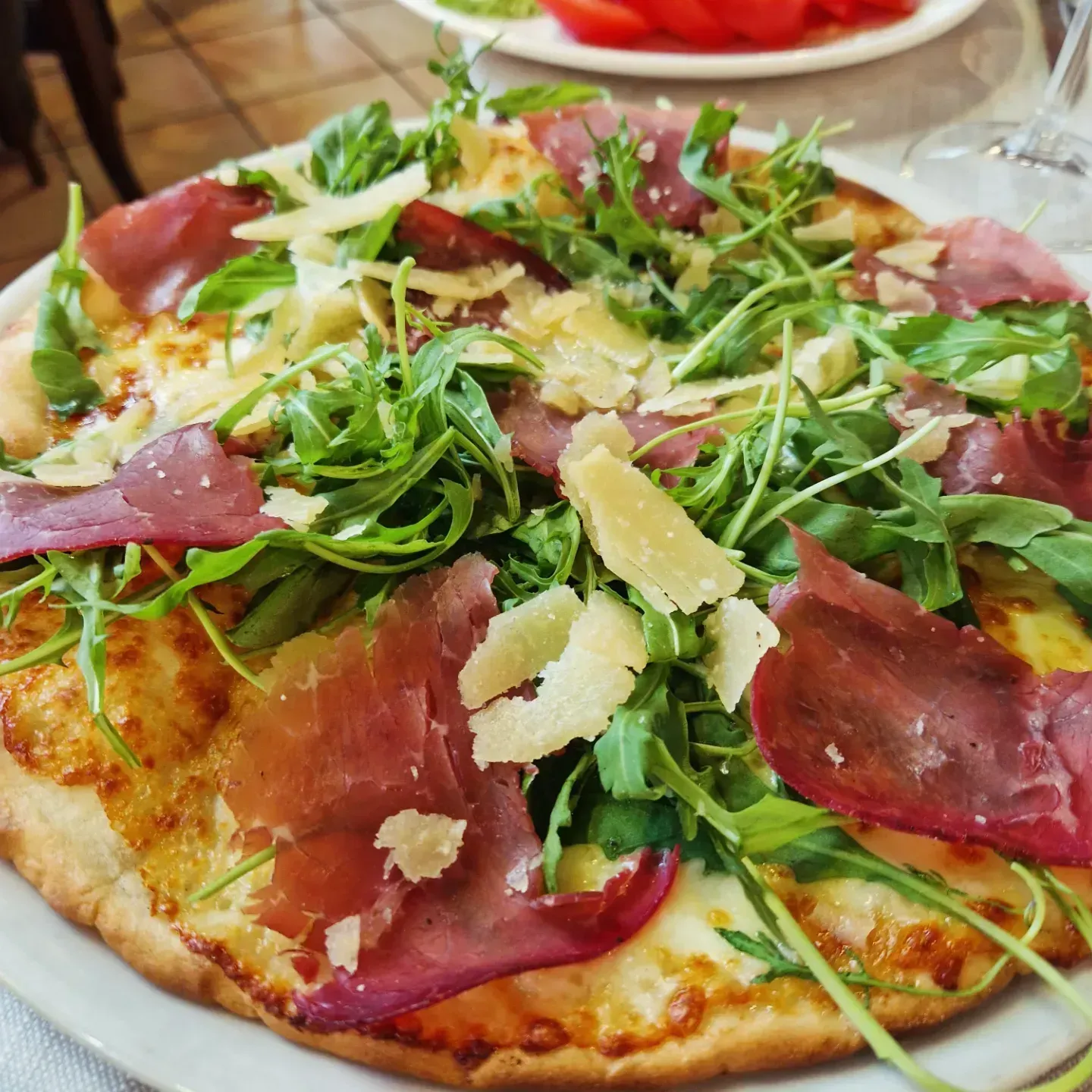 Pizza con rucola e prosciutto, guarnita con scaglie di formaggio e servita su un piatto bianco.
