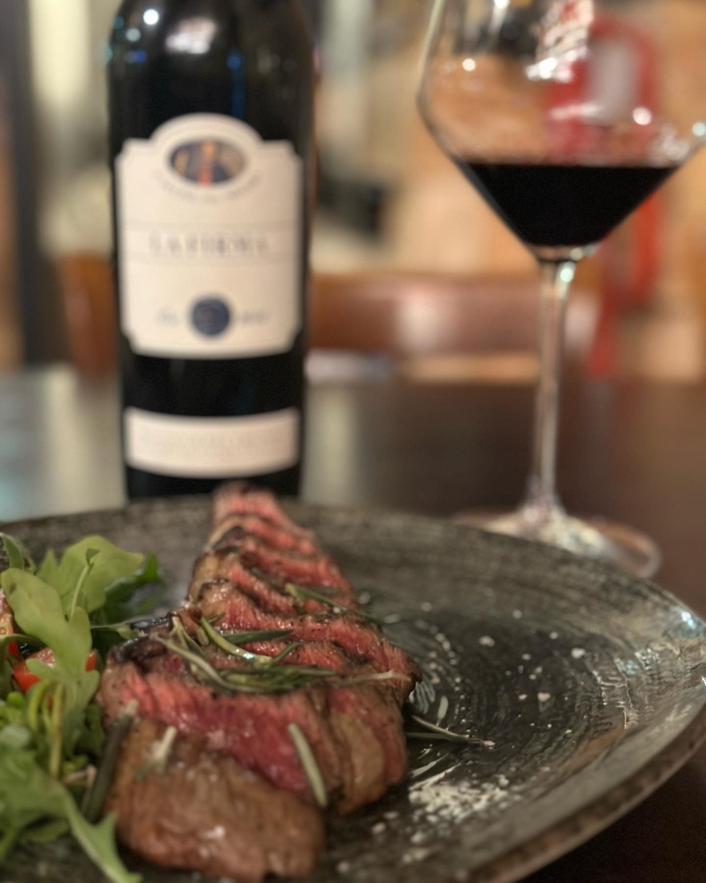 Cena a base di bistecca servita su un piatto con verdure, davanti a una bottiglia di vino e un bicchiere di vino rosso.