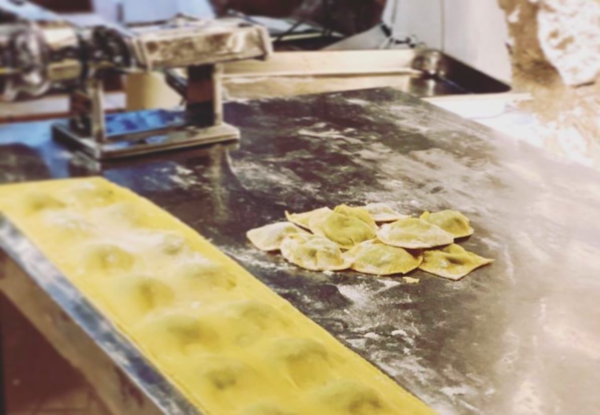 Mani che preparano ravioli su un tavolo da lavoro infarinato, con un vassoio giallo in primo piano.