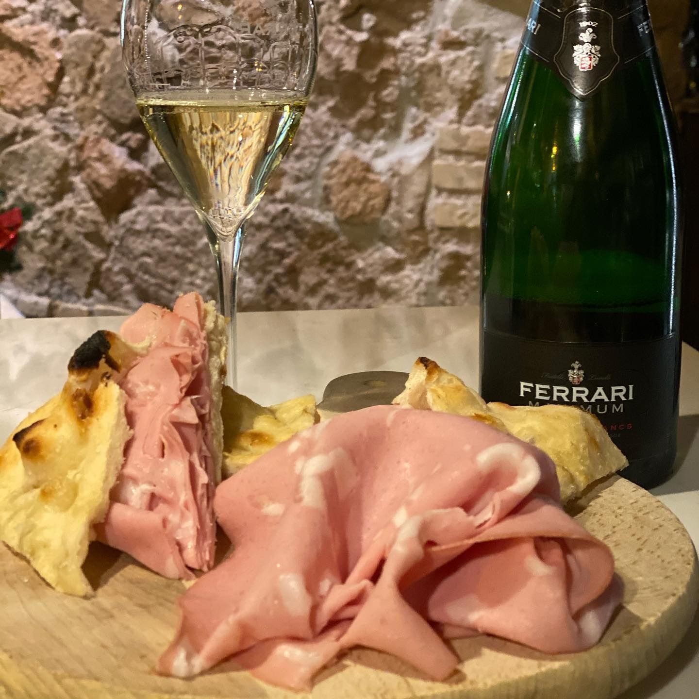 Un calice da vino, una bottiglia Ferrari e delle fette di prosciutto su un tagliere di legno appoggiato a un muro di pietra.